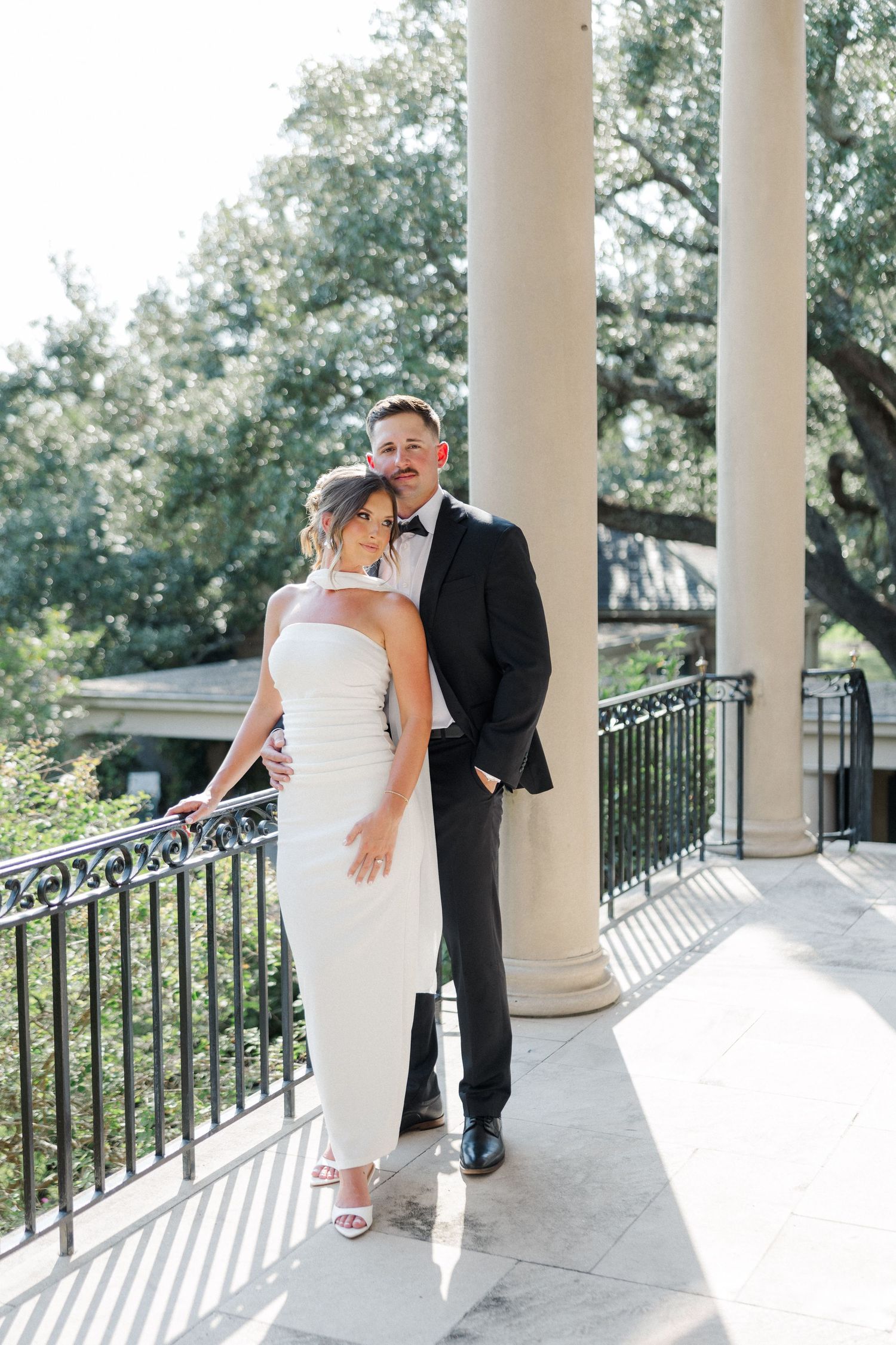 Elegant couple in wedding attire poses playfully between white columns on a porch with iron railings.