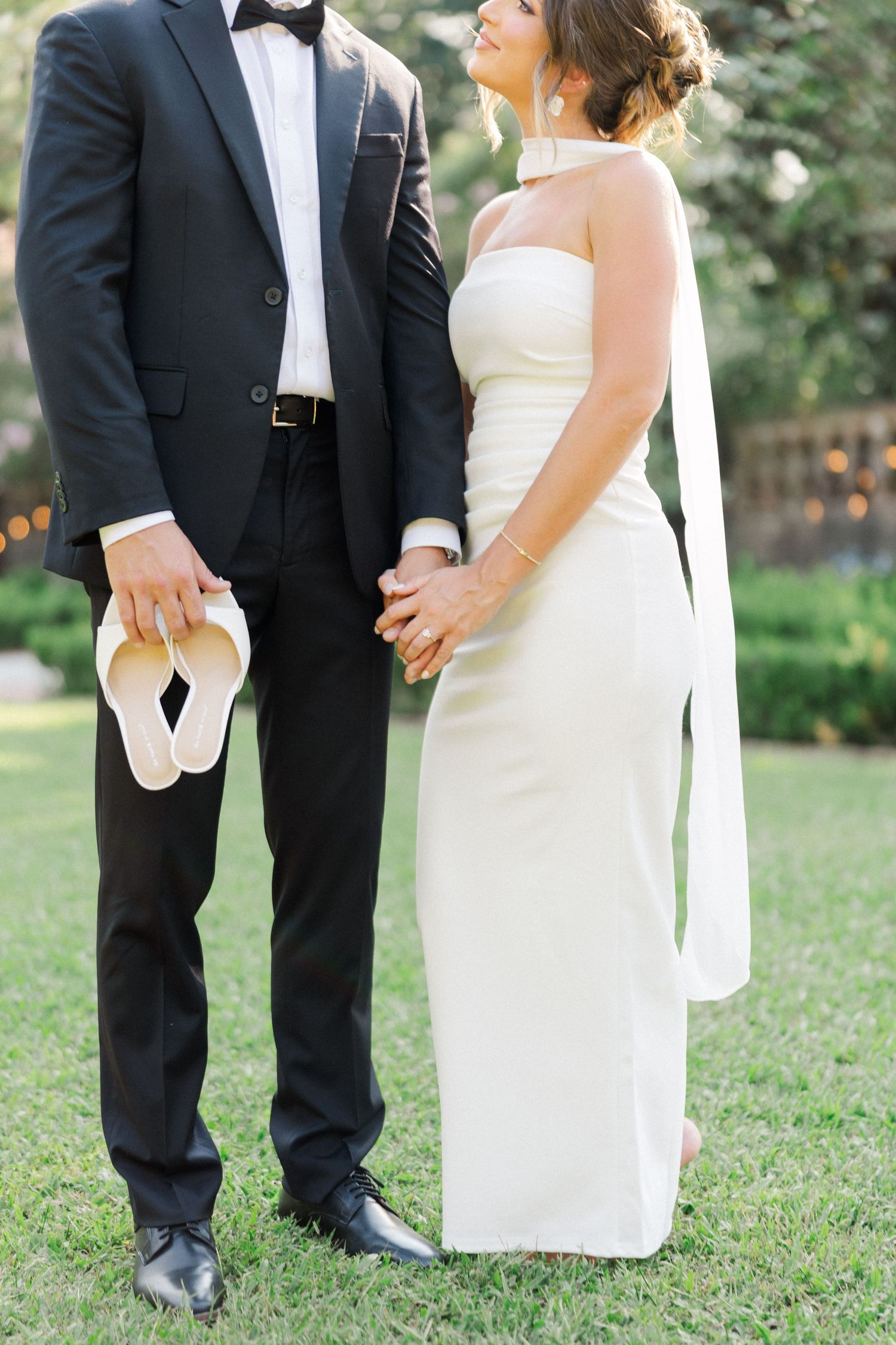 A couple in wedding attire stand together on grass, with the bride in white and groom in black tuxedo.