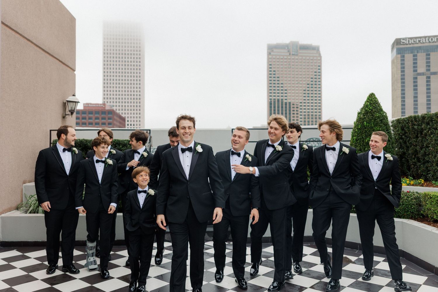 A group in formal black tuxedos poses together on a rooftop terrace with city buildings in the background.