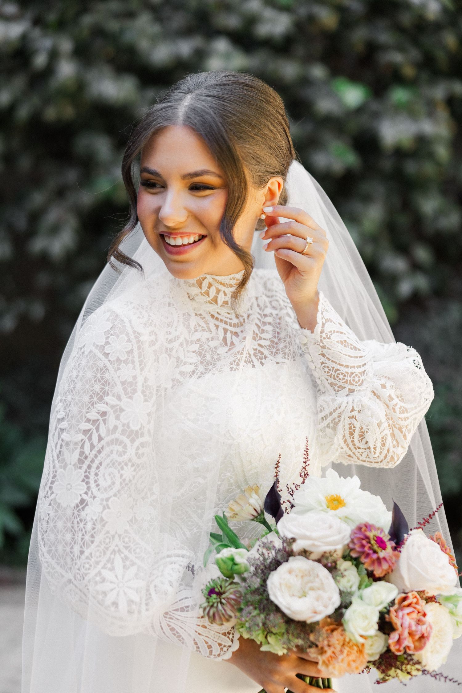 Bride in long sleeve lace wedding dress and veil holds colorful bouquet of flowers against blurred outdoor background.
