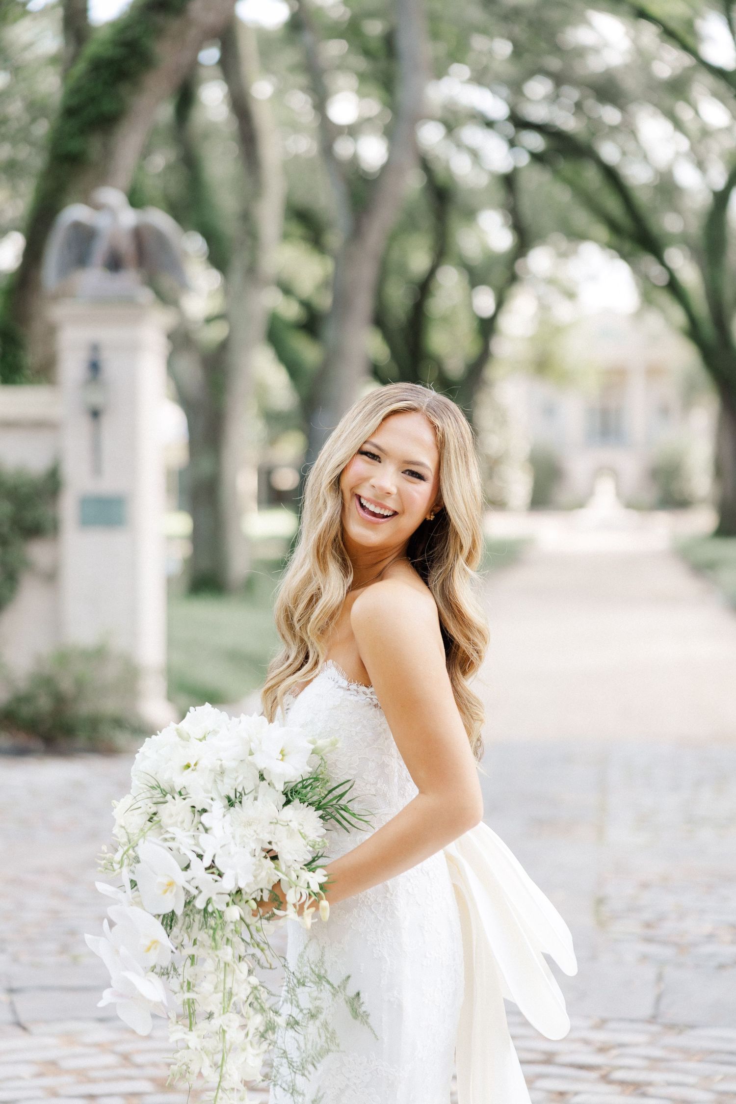 A bride in a white strapless wedding gown holds a lush white bouquet while standing on a tree-lined cobblestone path.