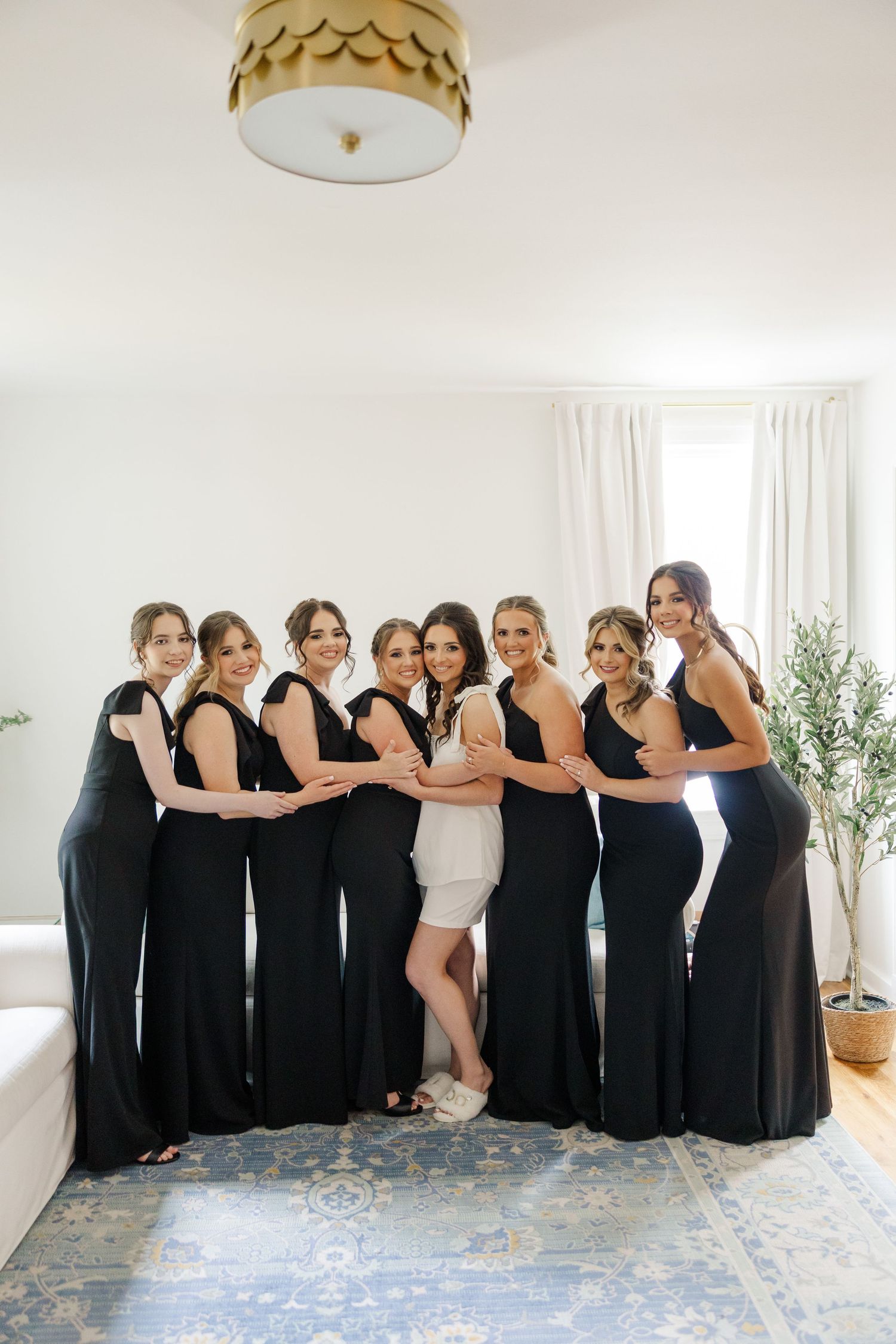 Bridesmaids in elegant black dresses pose together in a bright white room with a decorative rug.