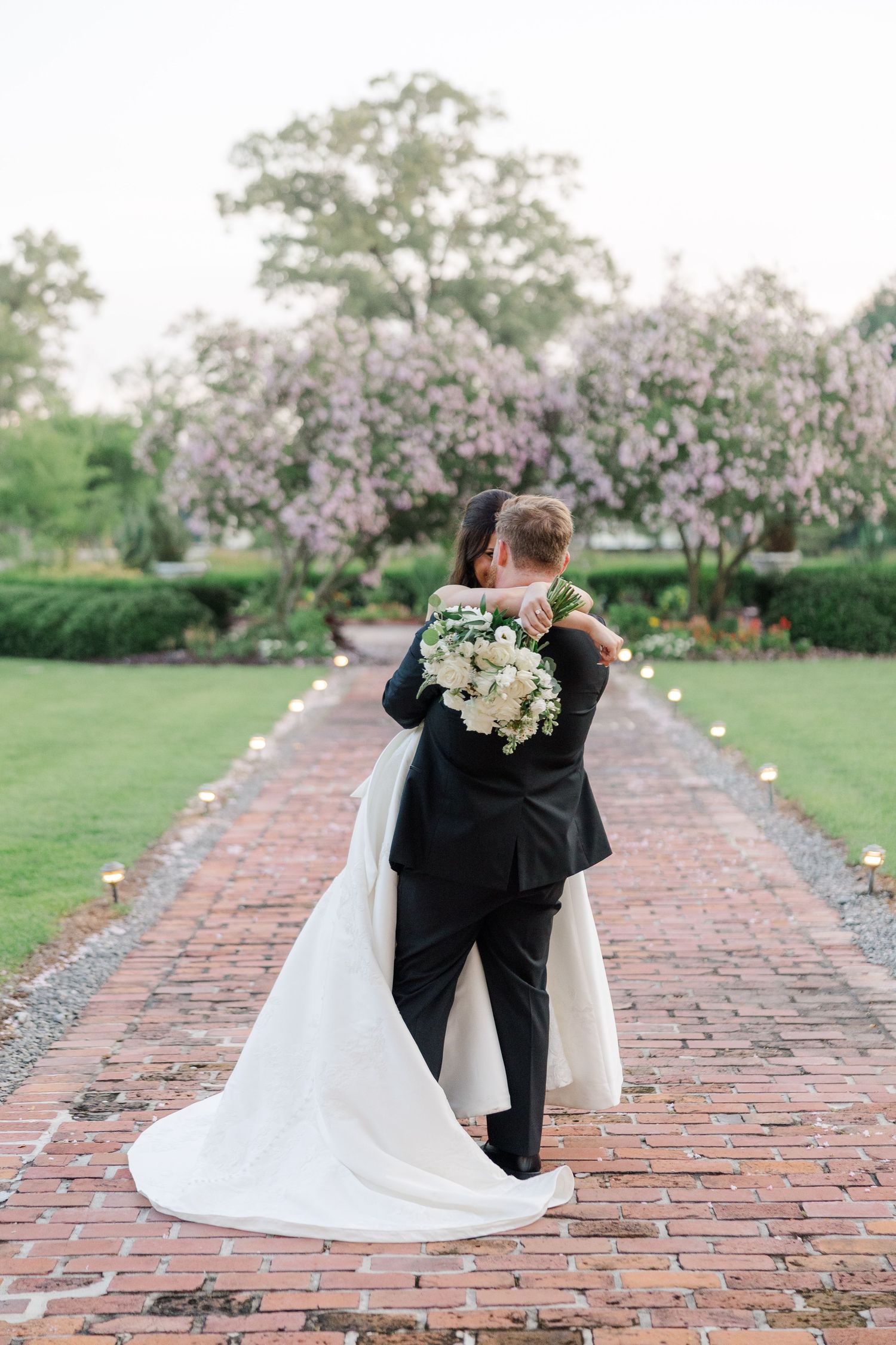 Wedding couple poses on brick pathway lined with blooming cherry blossom trees.