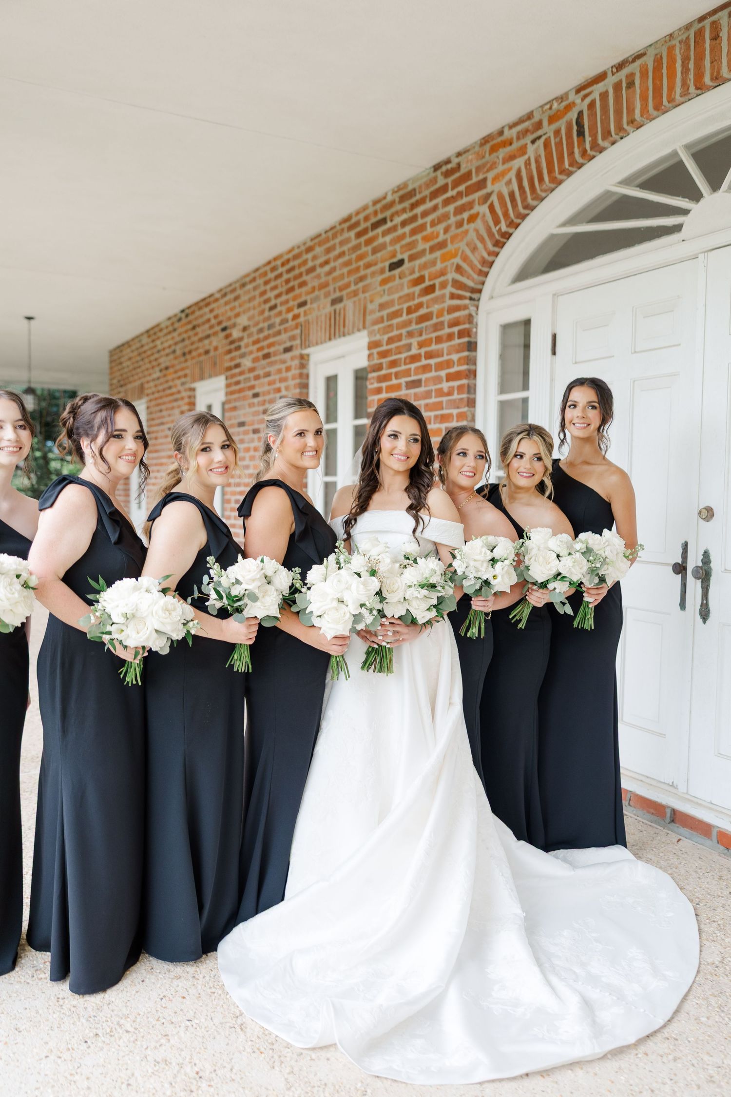 A bridal party in black dresses poses with the bride in white against a brick exterior wall.