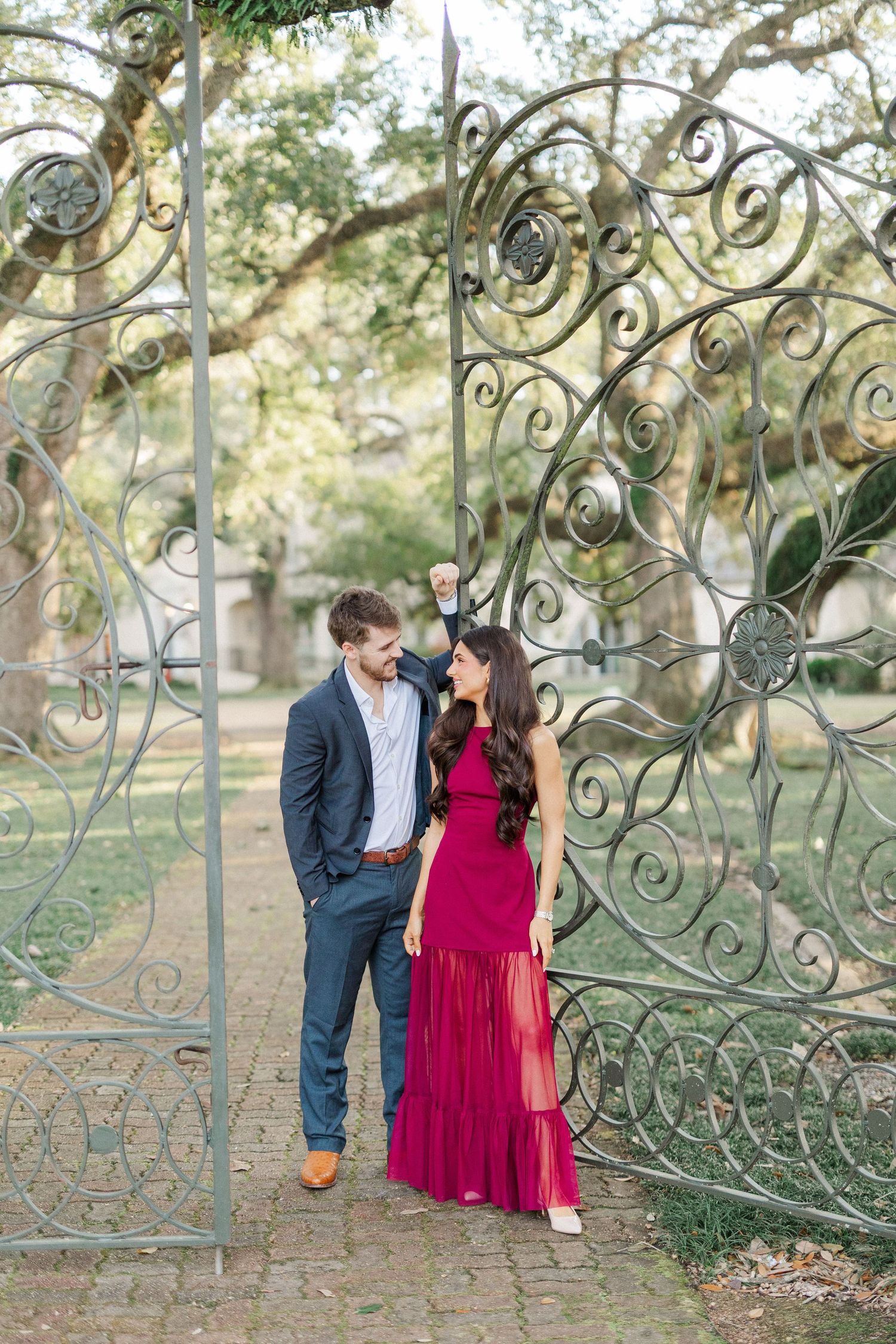 Couple poses together by decorative wrought iron garden gate with her in flowing red dress and him in navy suit.