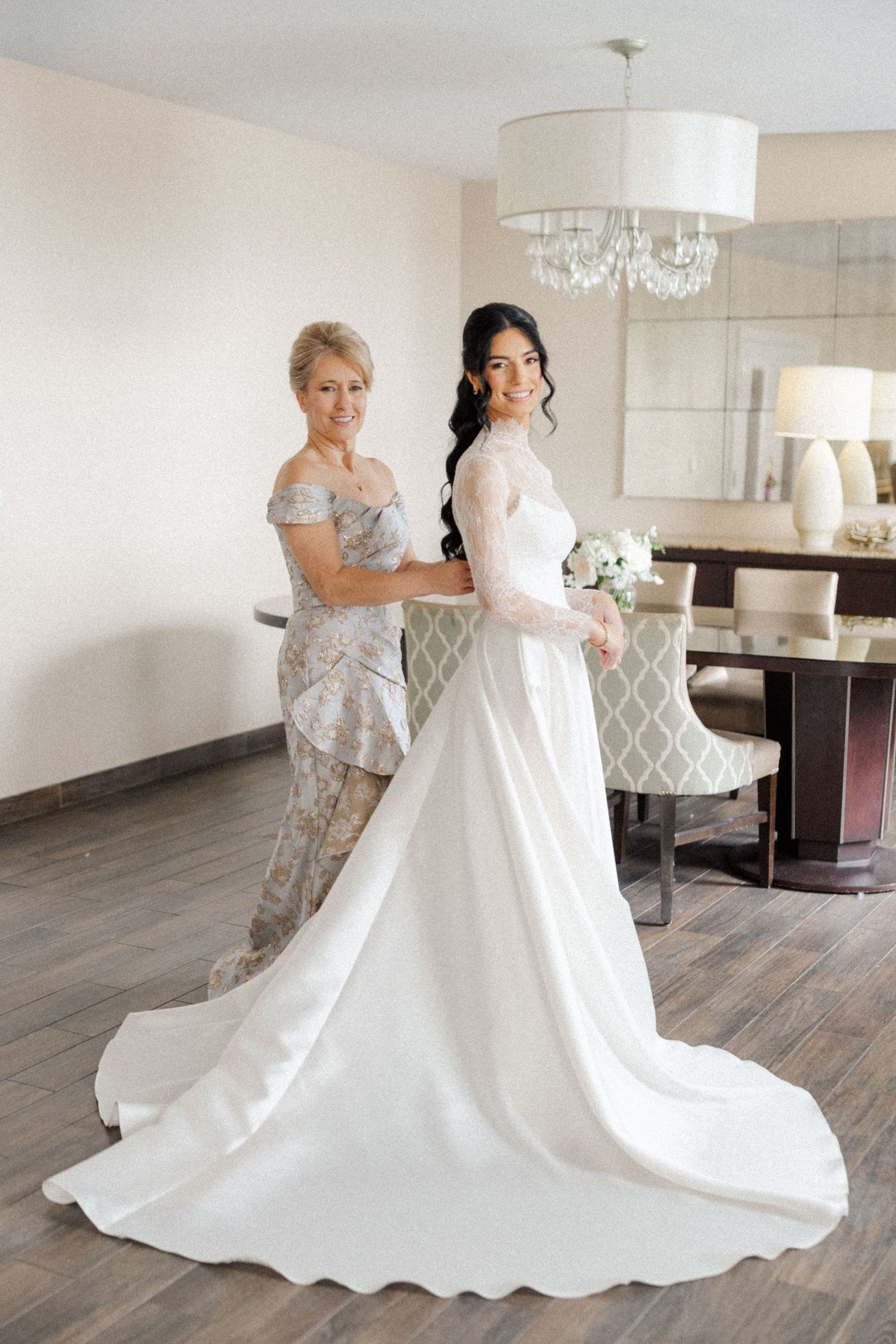 Mother helps bride in long white gown with train prepare in a bright hotel suite.