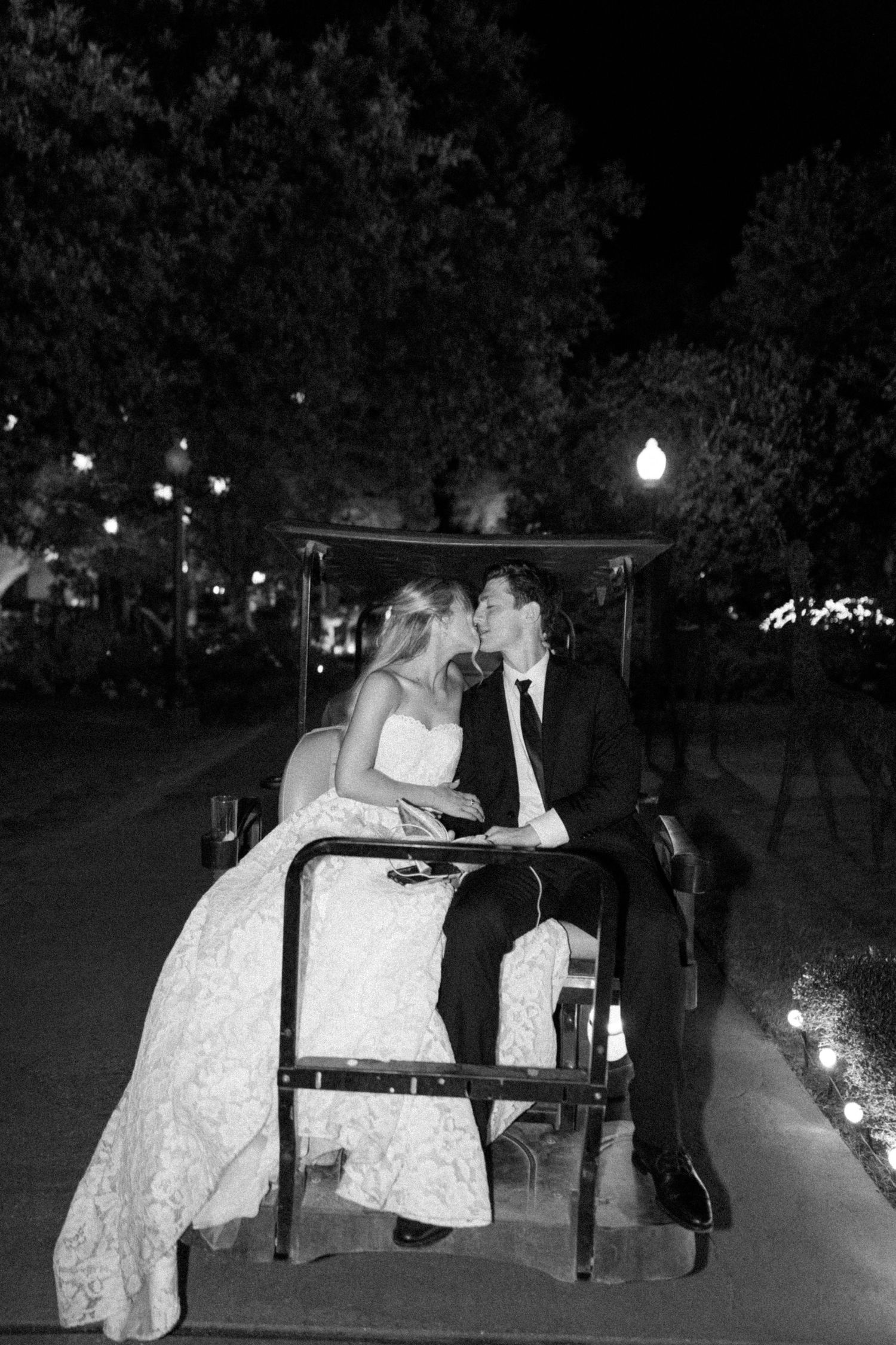 Black and white photo of bride and groom kissing on golf cart under evening lights after wedding reception.