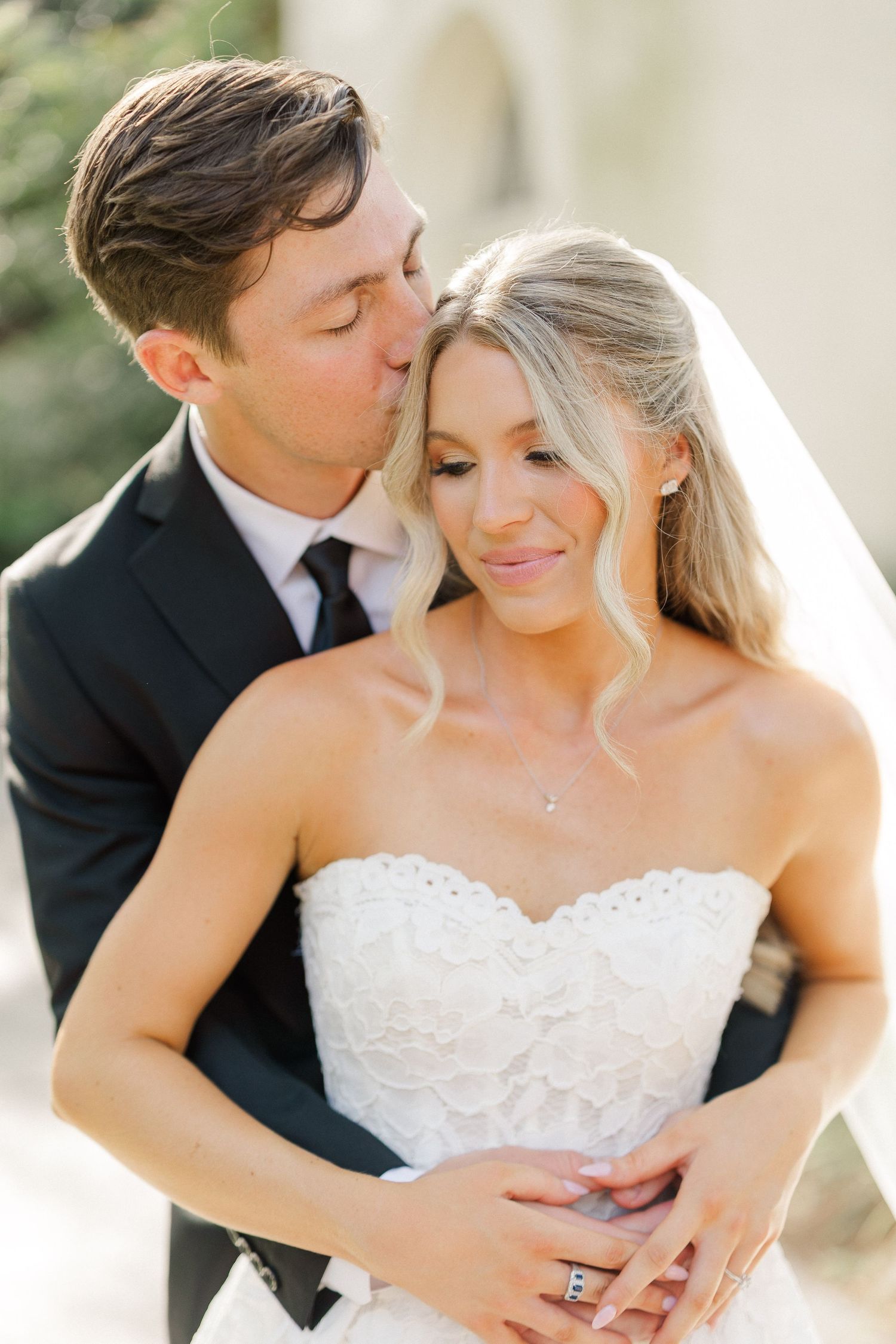 Groom tenderly kisses blonde bride wearing a strapless lace wedding gown and delicate veil outdoors.