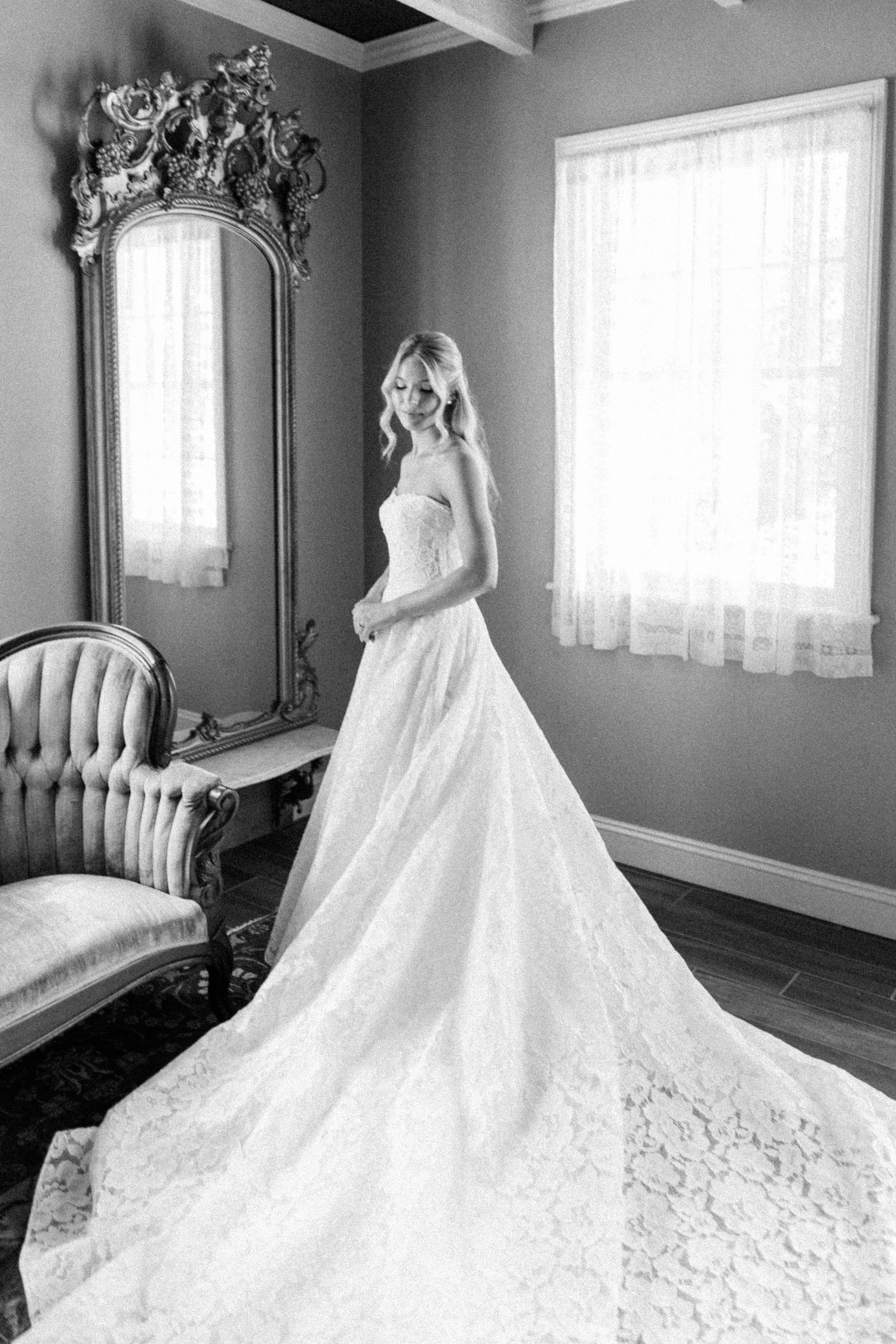 Bride in strapless lace ballgown wedding dress poses before ornate mirror in elegant black and white bridal portrait.