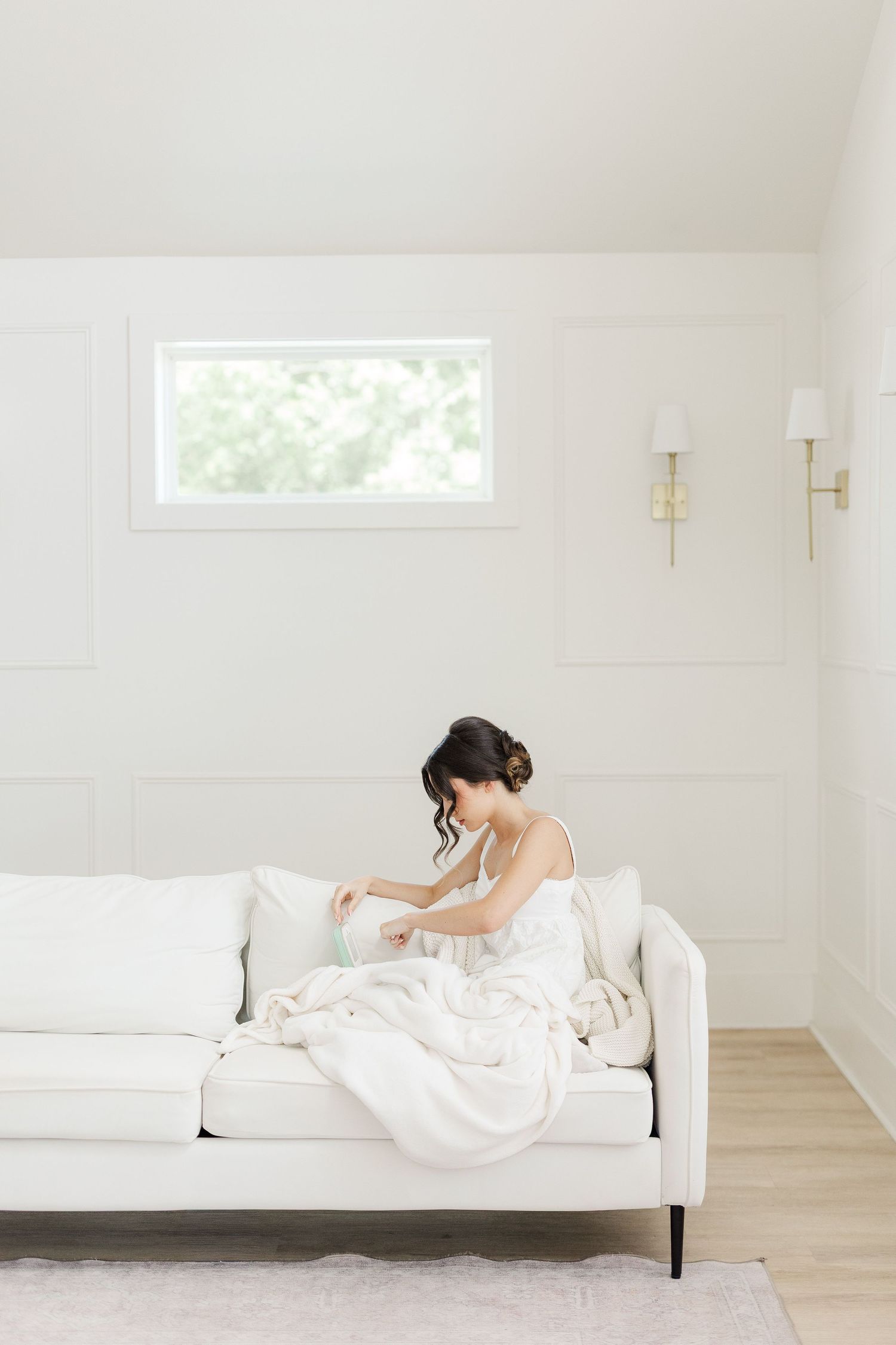 Bride in white gown sitting on ivory sofa adjusting her dress in a bright, minimalist bridal suite.