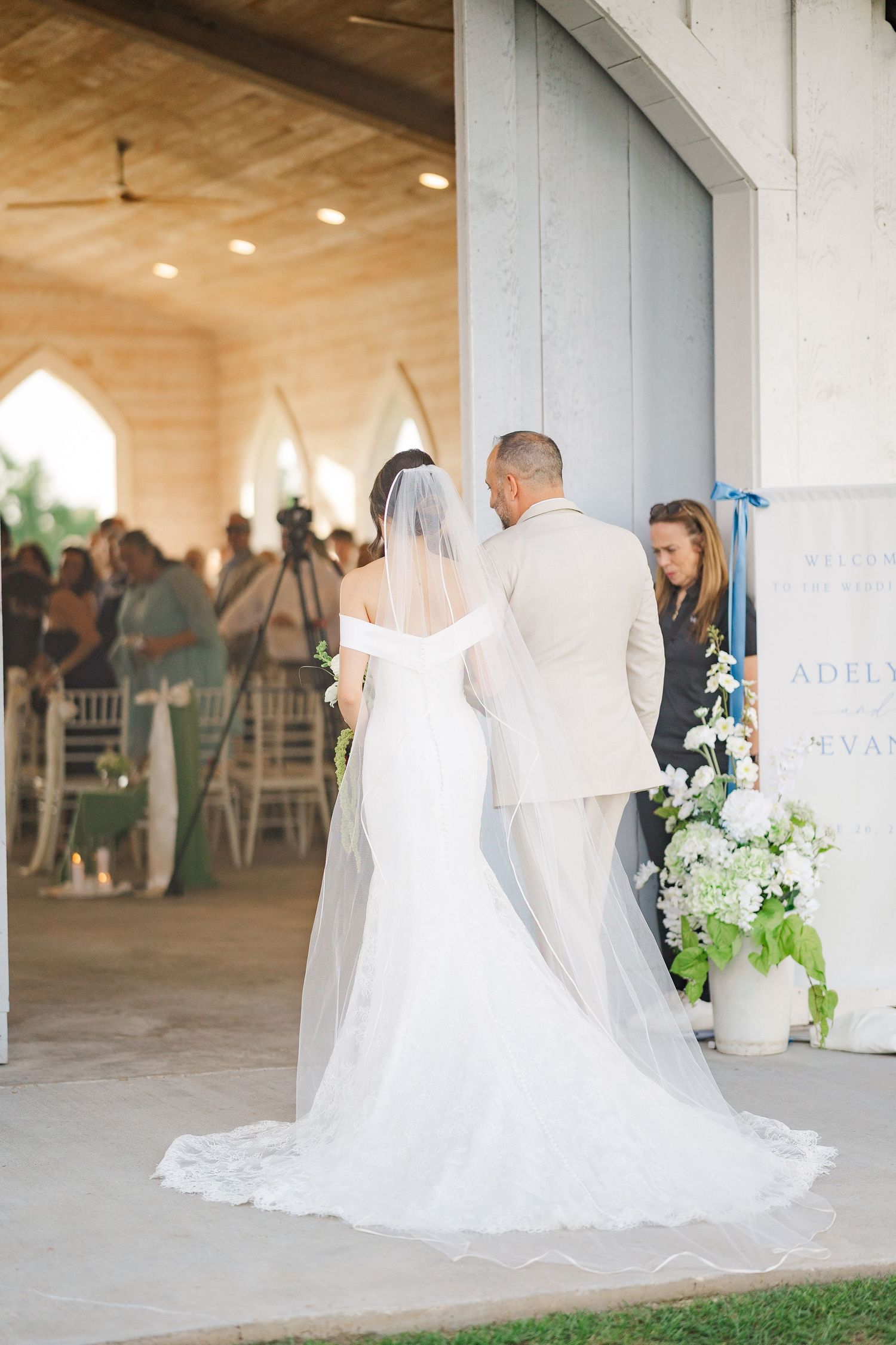 Bride in off-shoulder gown and veil walks down the aisle with escort at an elegant outdoor ceremony.