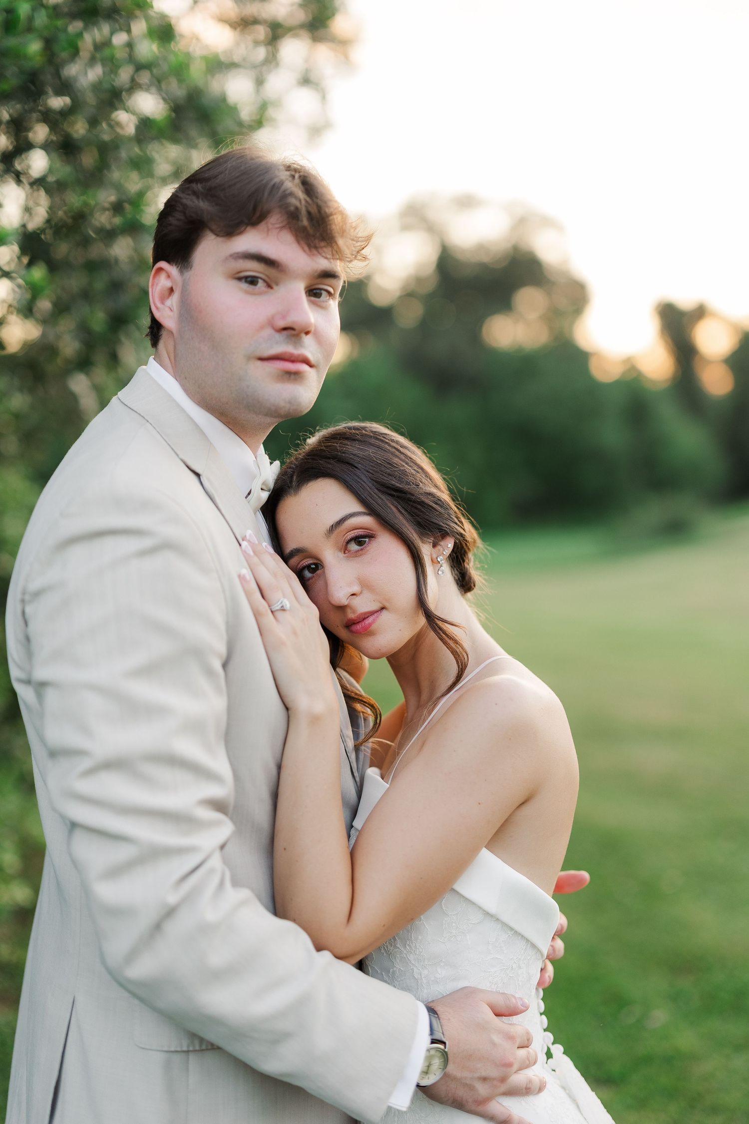 Bride and groom embrace outdoors at golden hour, she in white gown, he in beige suit, green trees behind.