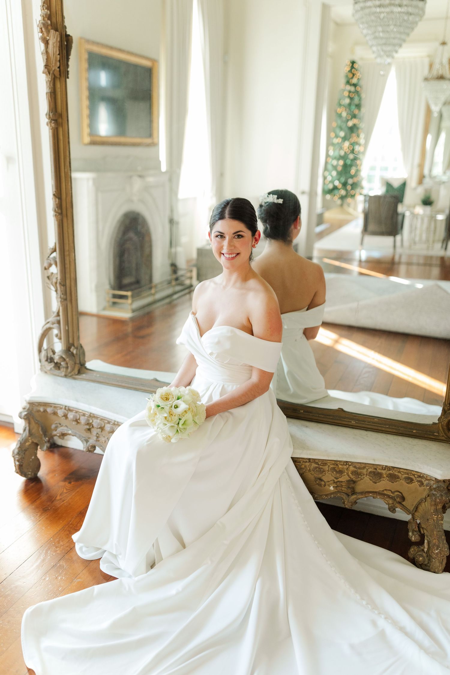Bride in strapless gown sitting on antique bench with reflection in gilded mirror holding white bouquet.