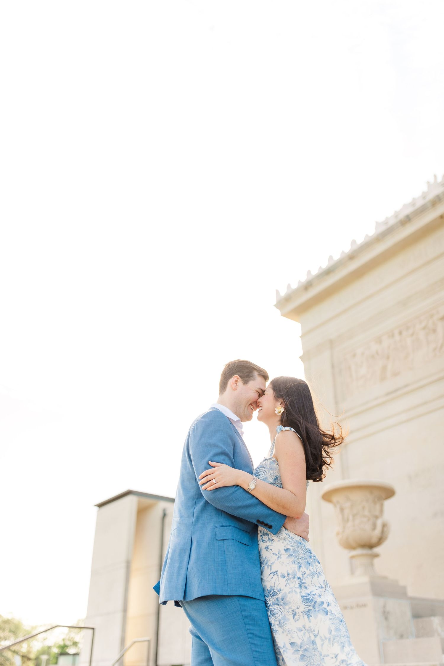 A romantic couple embraces in formal attire against an elegant stone building backdrop in soft lighting.