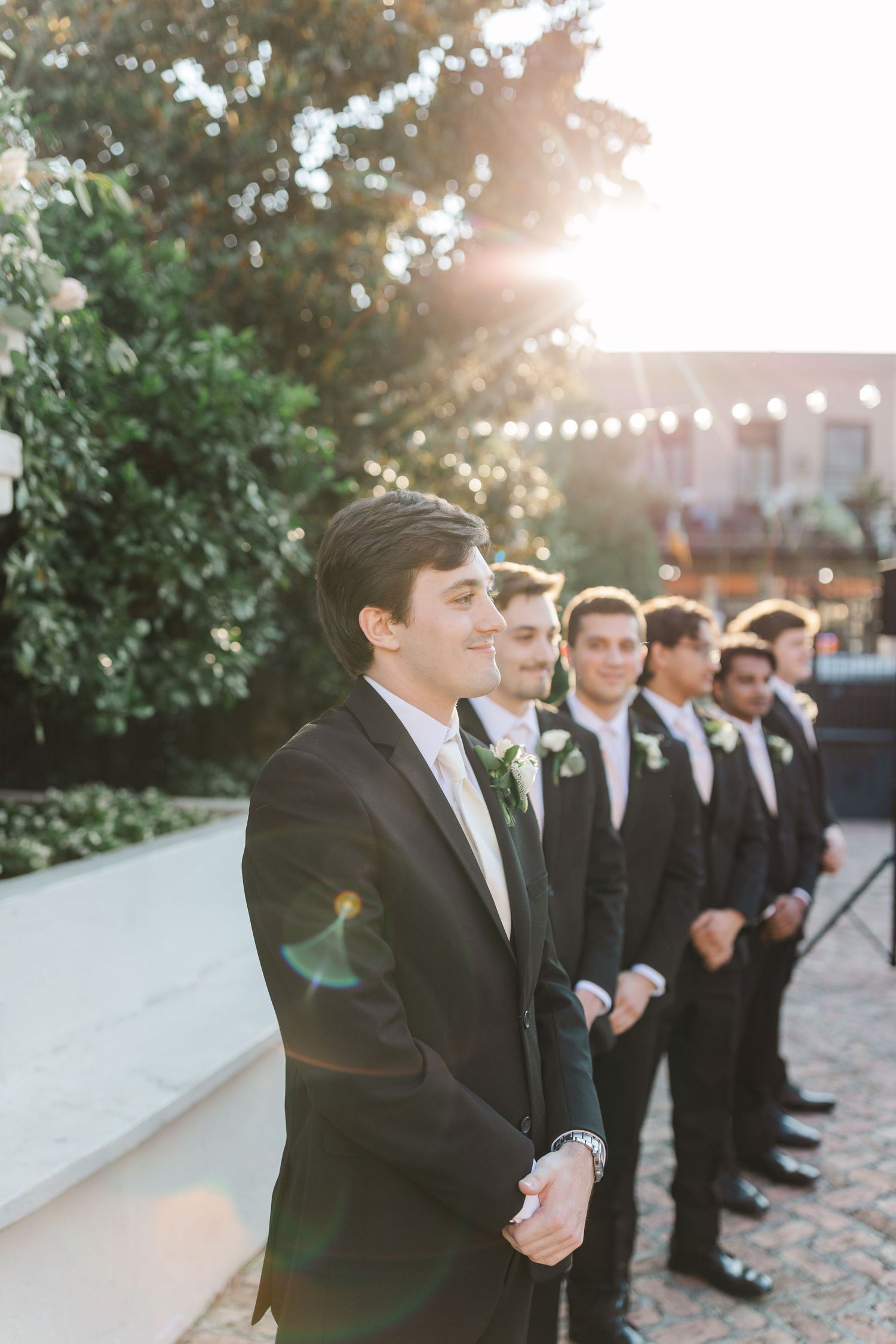 Groomsmen stand in line during golden hour at outdoor wedding venue.