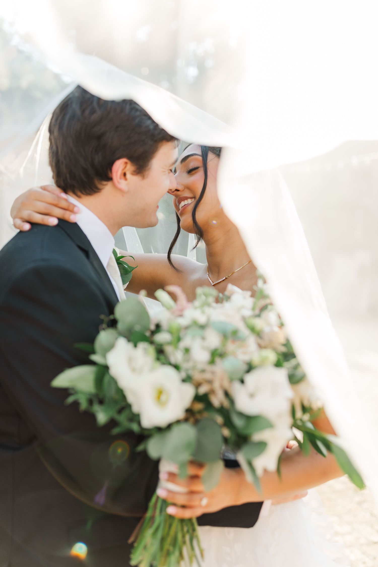 Joyful wedding couple sharing intimate moment with white floral bouquet at outdoor venue.