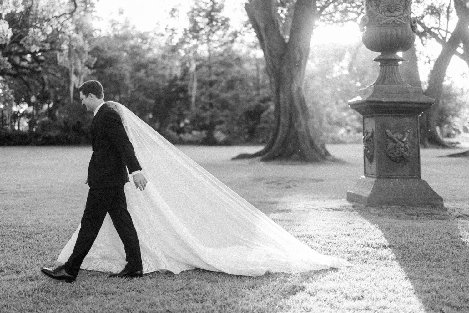 Groom in black tuxedo walks beside flowing bridal veil train on lush garden lawn, black and white.