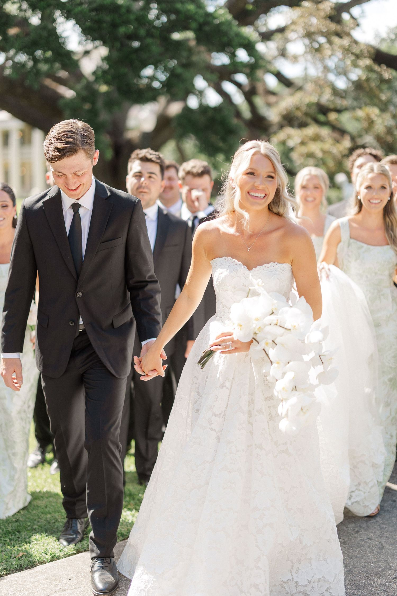 Smiling bride in strapless lace gown holds white orchid bouquet while walking with groom in black suit outdoors.
