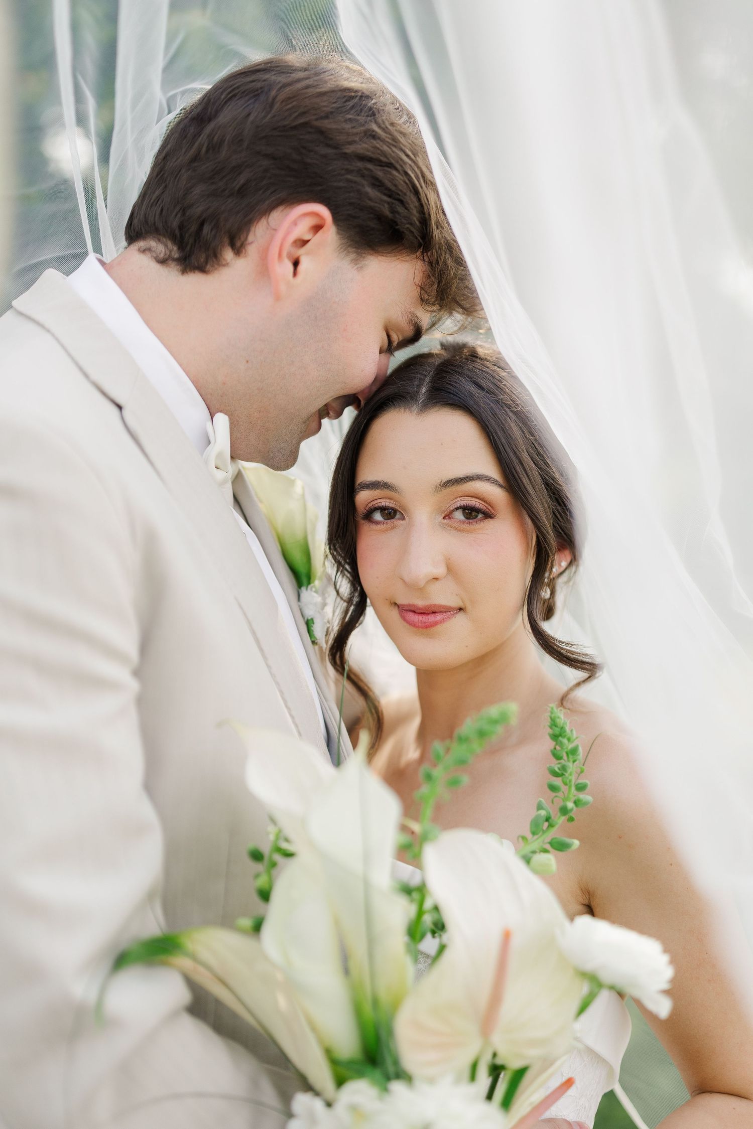 Groom in cream suit tenderly nuzzling bride holding white floral bouquet beneath a flowing veil.