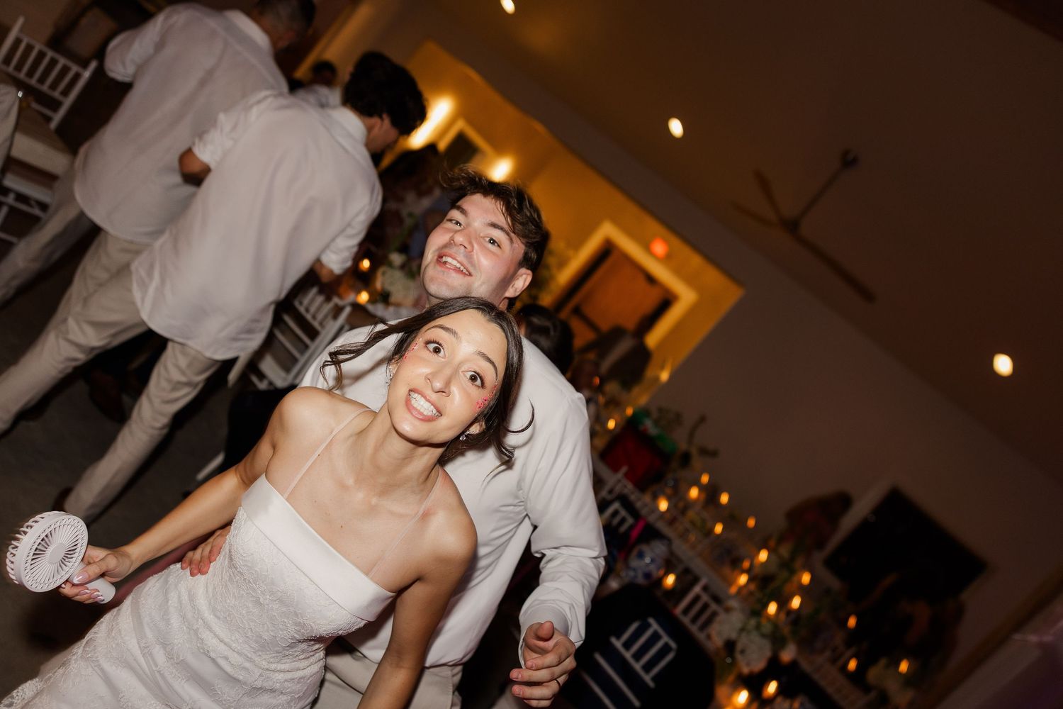 Smiling bride and groom dance together at wedding reception, surrounded by guests and warm ambient lighting.