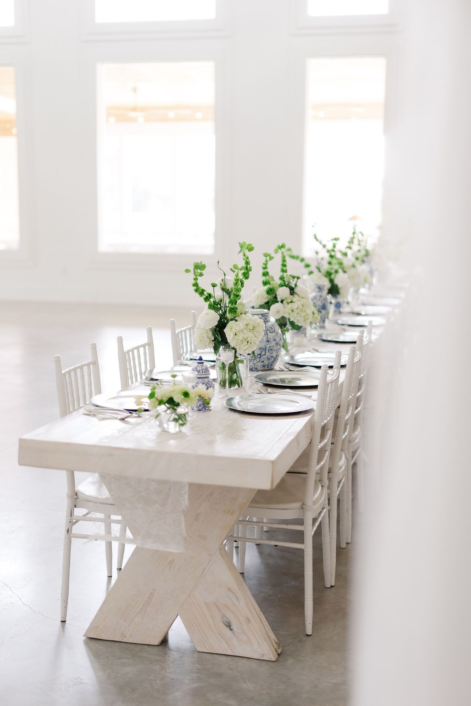 Elegant white farmhouse table with blue and white floral centerpieces set for a wedding reception in a bright white venue.