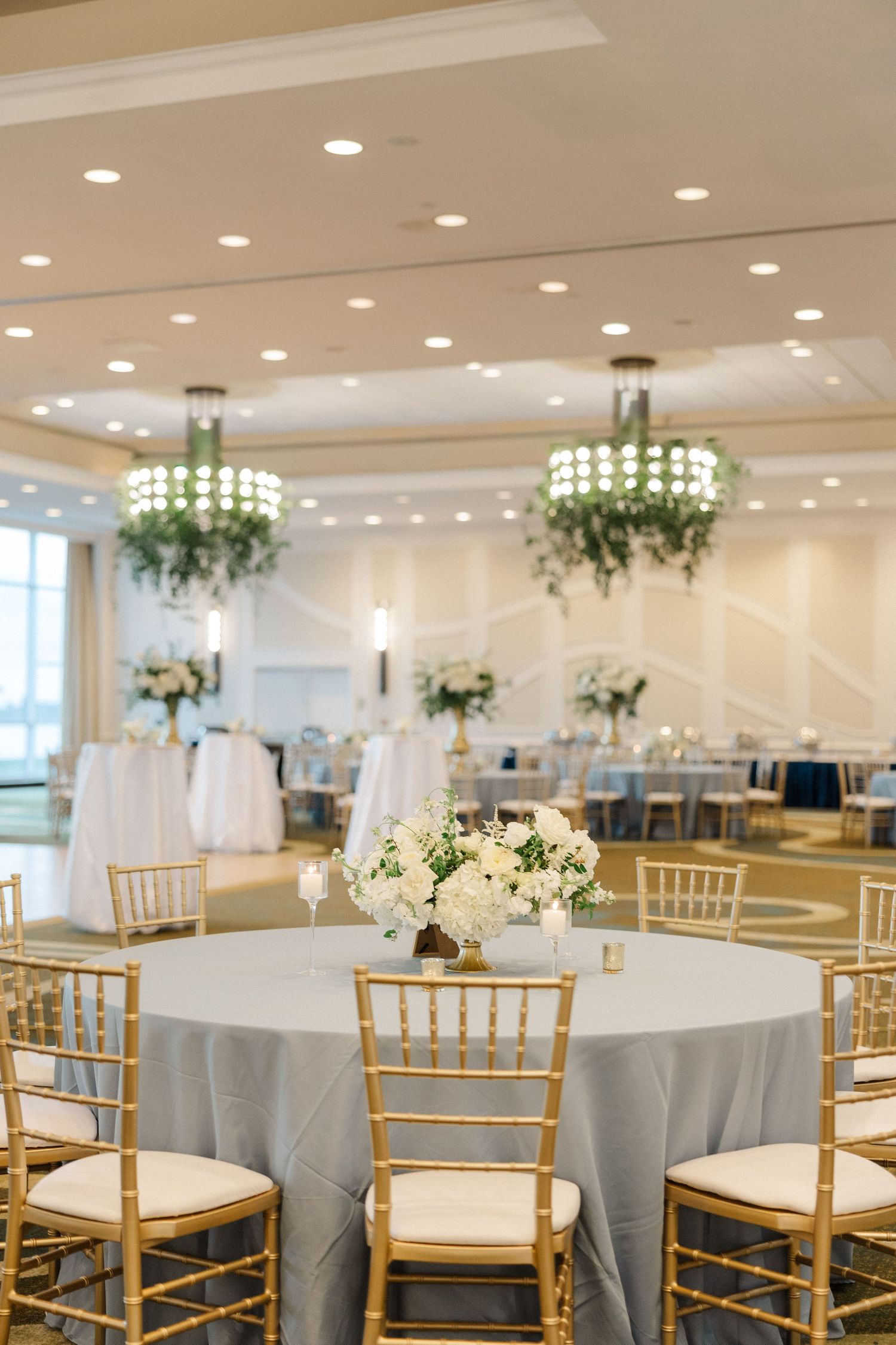 Elegant wedding reception ballroom with gold chiavari chairs, gray linens, white floral centerpieces, and hanging greenery chandeliers.
