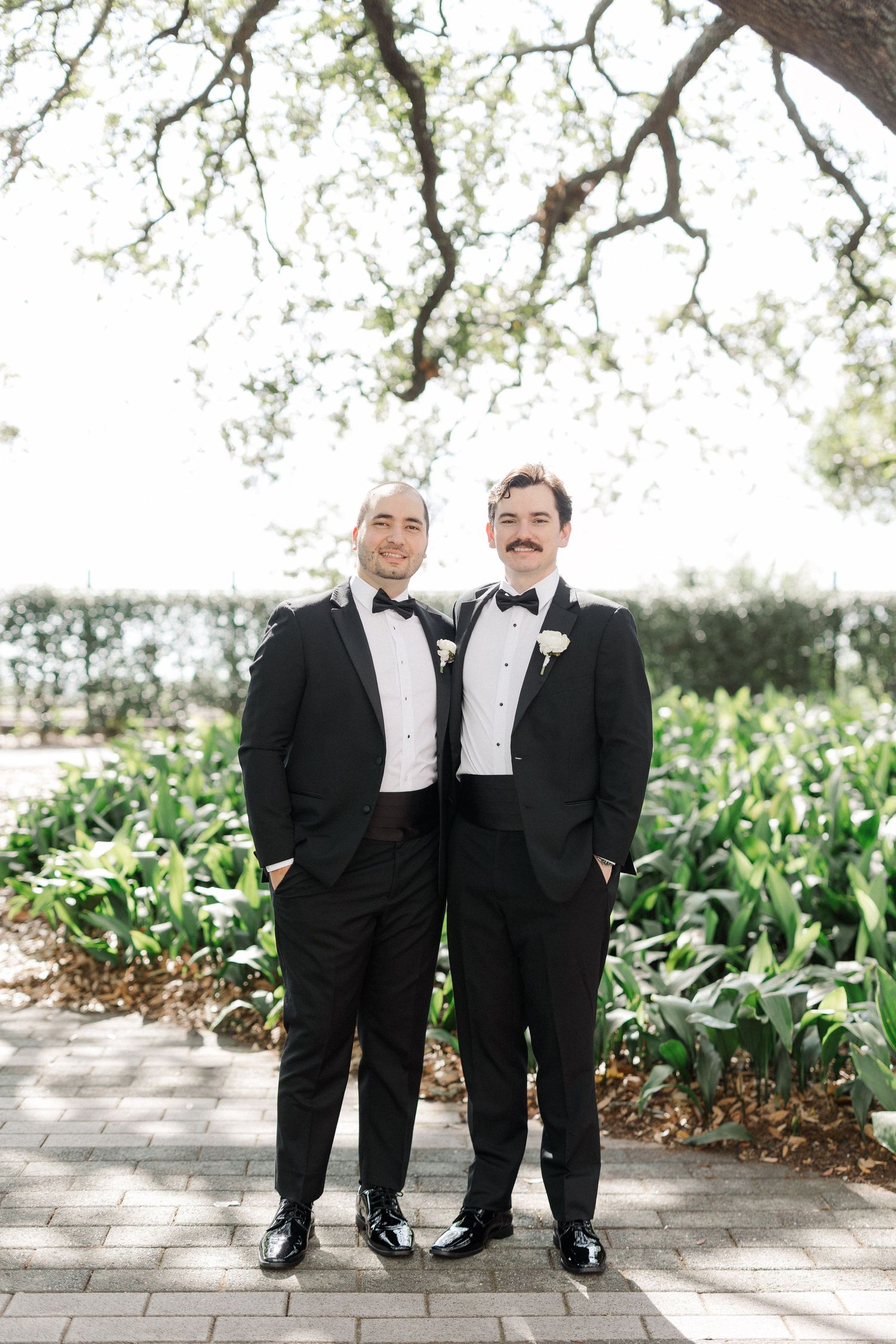 Two grooms in black tuxedos with bow ties and boutonnieres smile together outdoors on their wedding day.