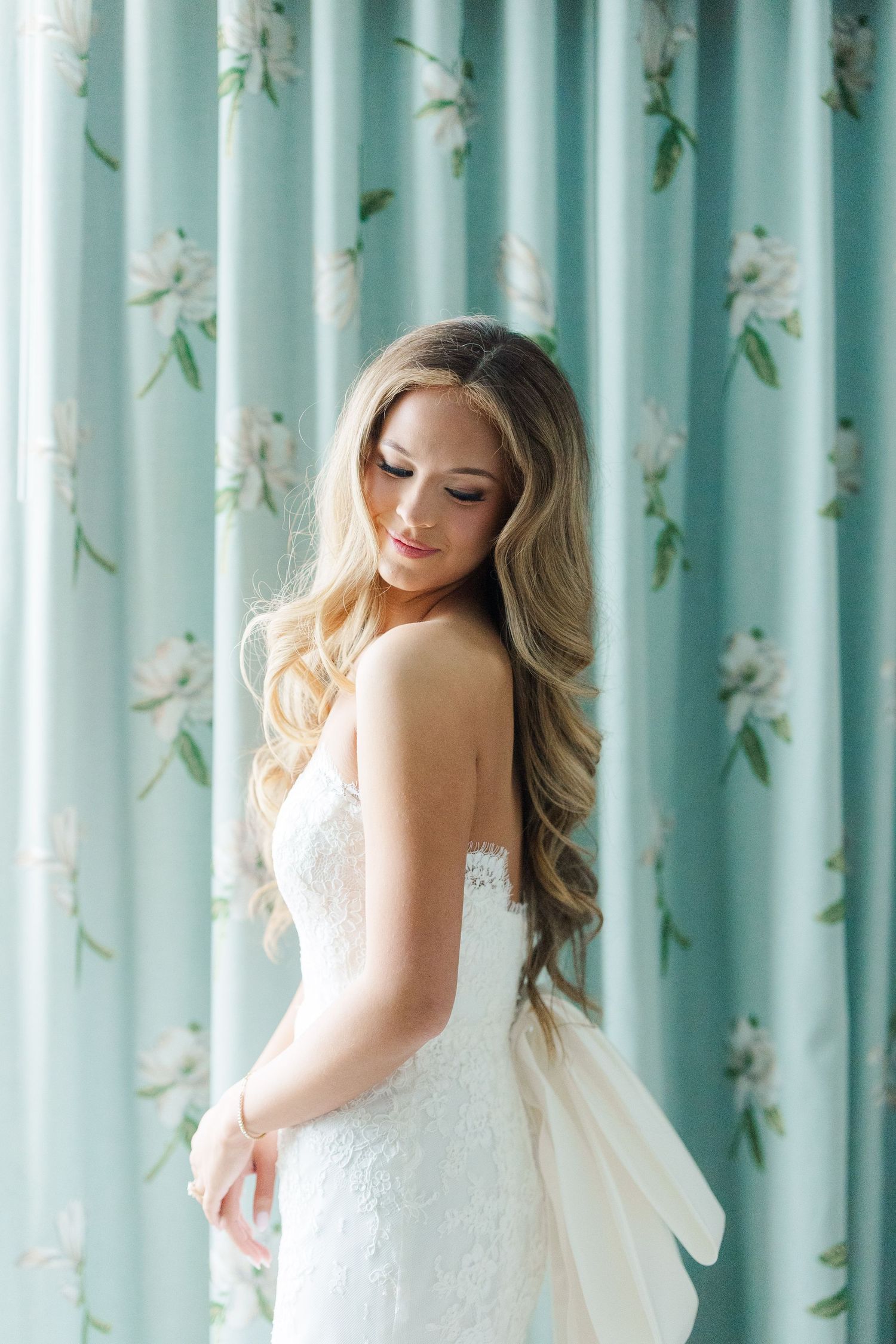 Elegant white lace wedding dress shown against mint floral curtains in soft natural light.