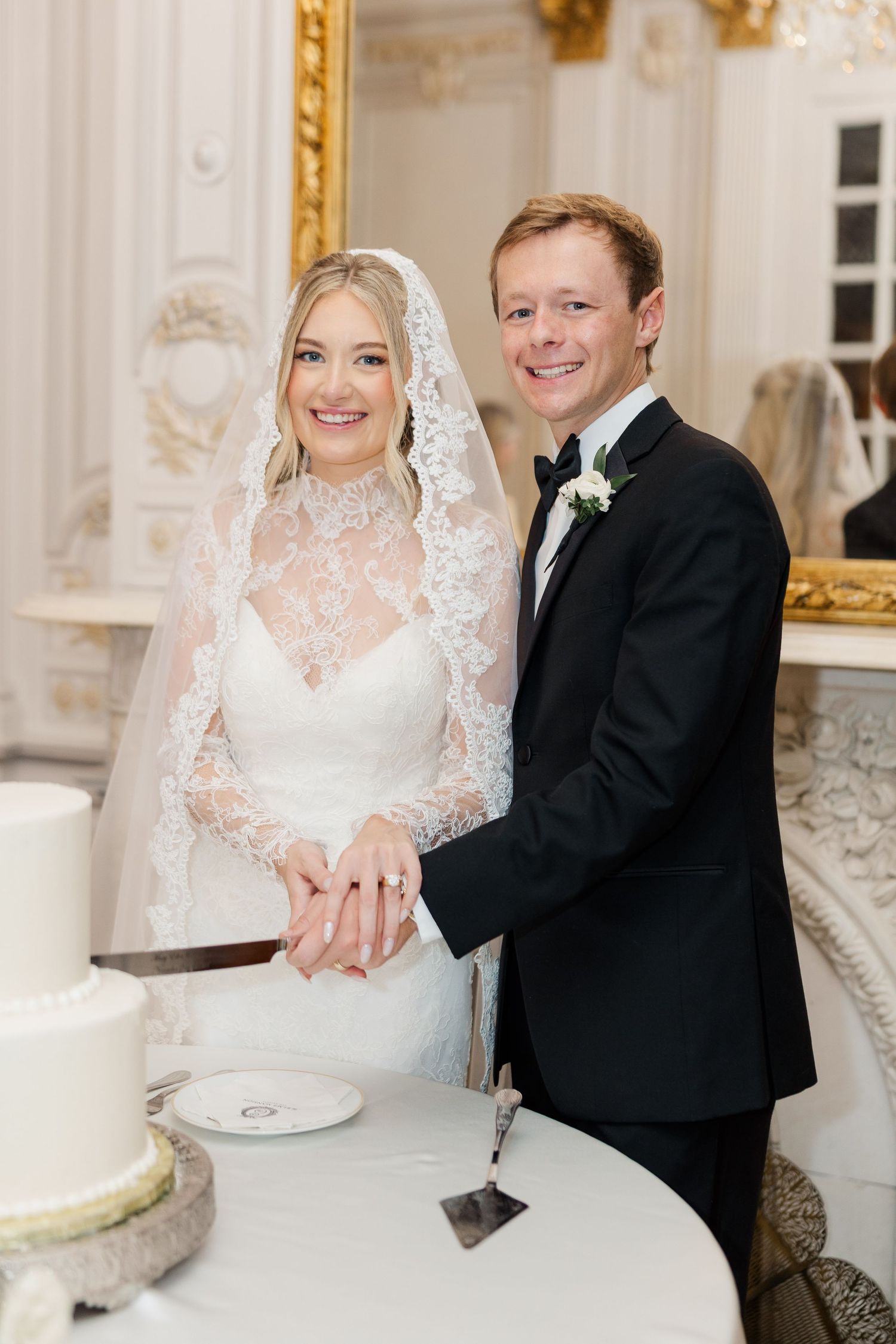 Wedding cake cutting moment in an ornate room with gold-framed mirrors and white decor.