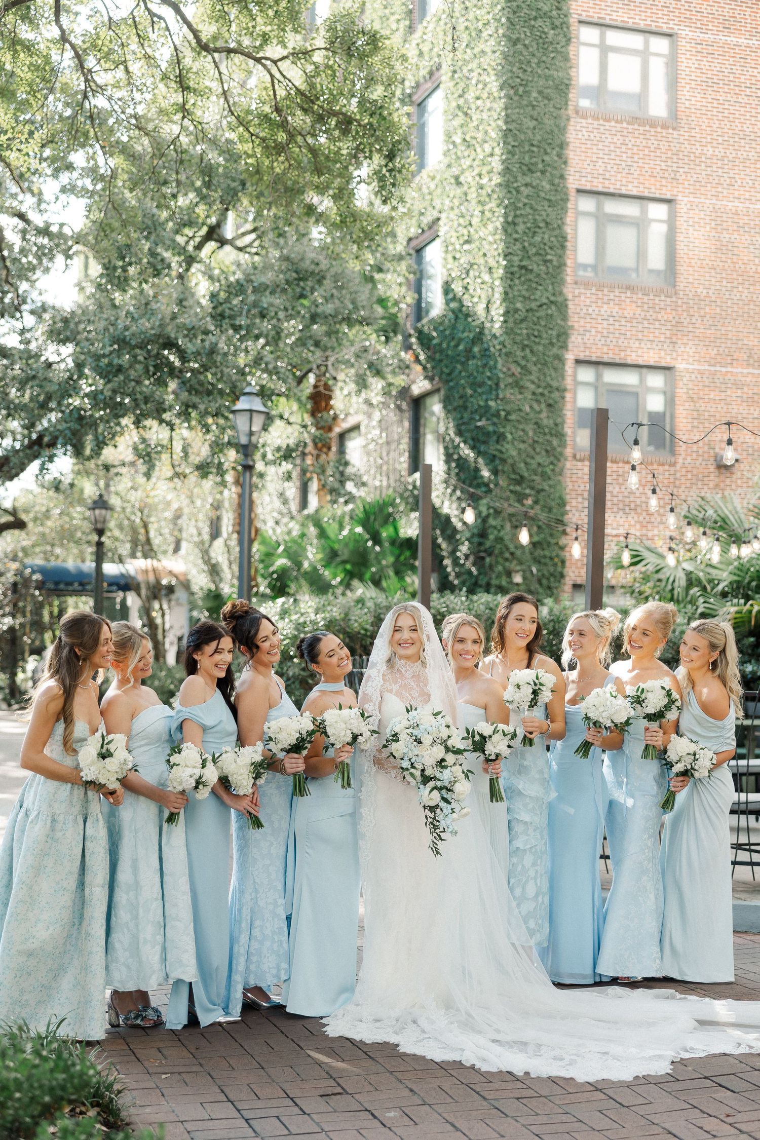 Bridal party in light blue dresses poses together on brick paved courtyard.