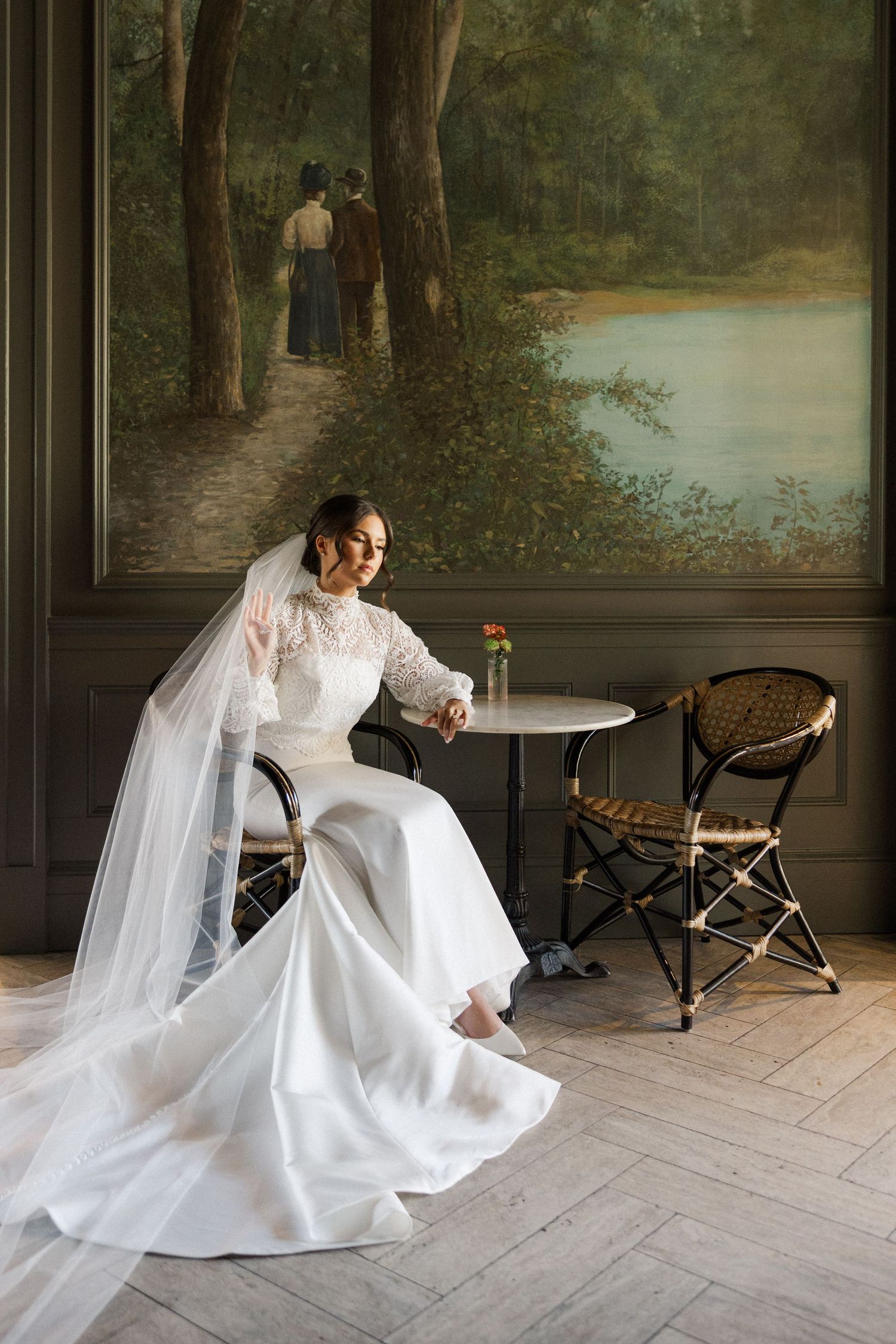 Elegant bridal portrait in white gown and veil seated at cafe table with romantic forest mural backdrop.