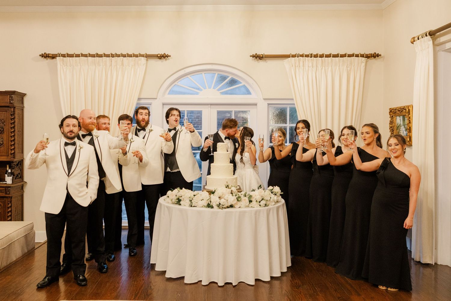 Wedding party in black dresses and white tuxedos poses together around a cake table.