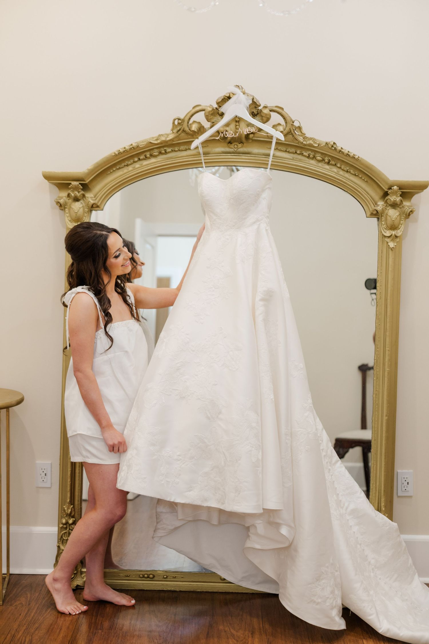 White wedding dress hanging in front of ornate gold mirror during bridal preparation.