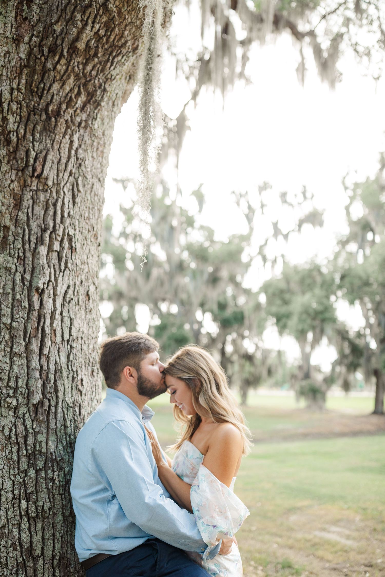 A romantic couple embraces beneath a Spanish moss draped oak tree in a dreamy outdoor setting.