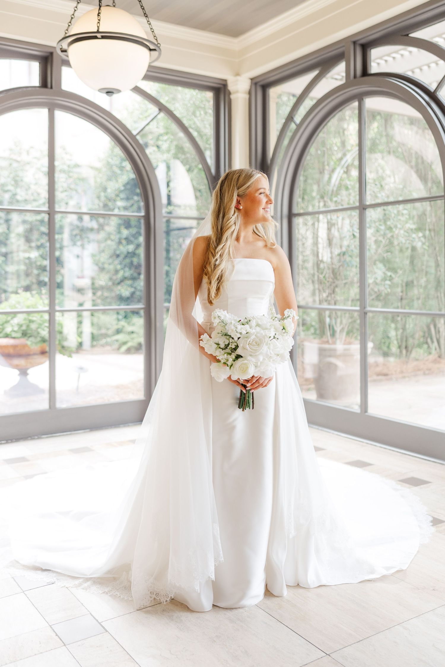 A white wedding dress and veil are captured in a beautiful light-filled room with arched windows.