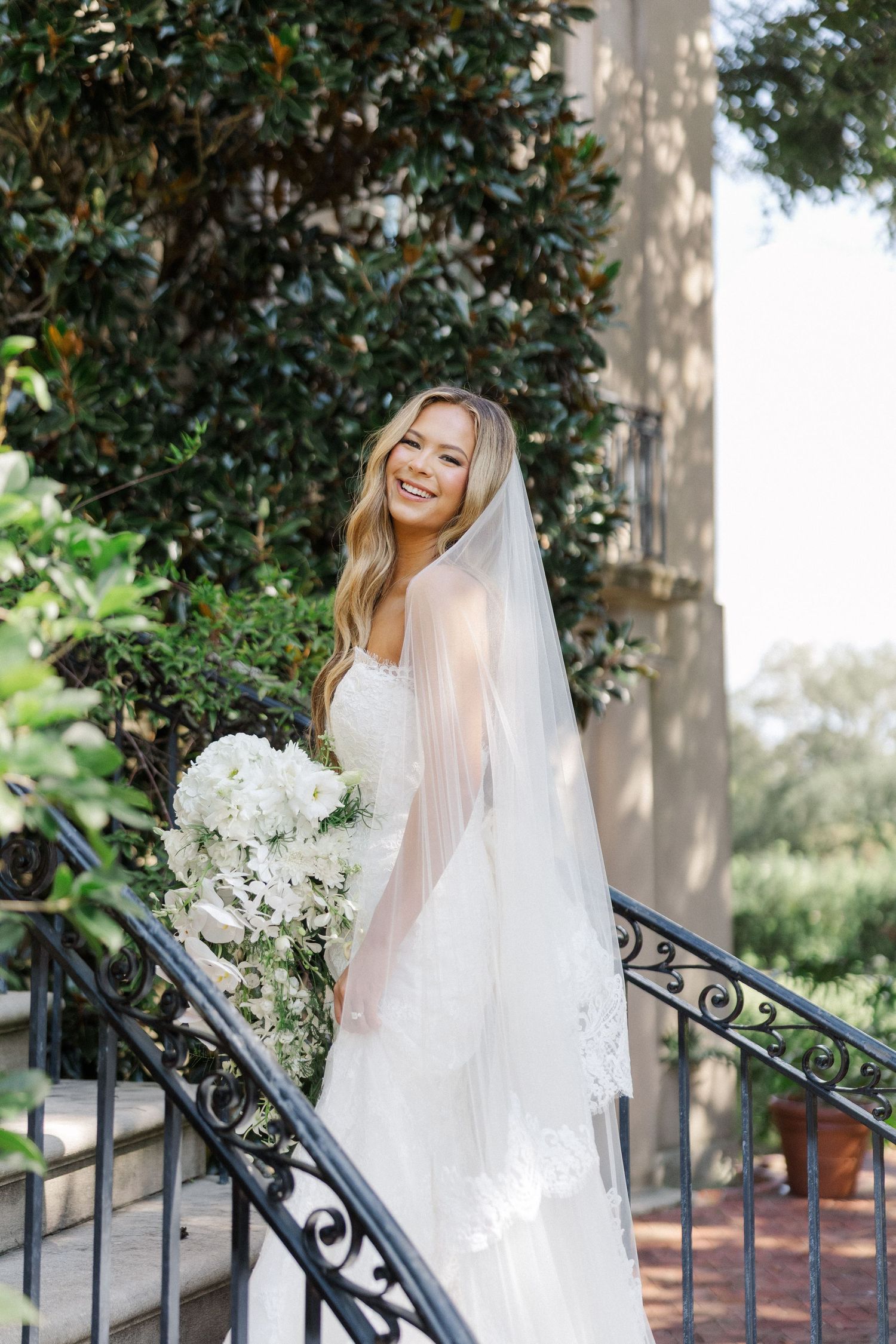 A bride in a flowing white gown and veil poses elegantly on an ornate staircase with white floral bouquet.