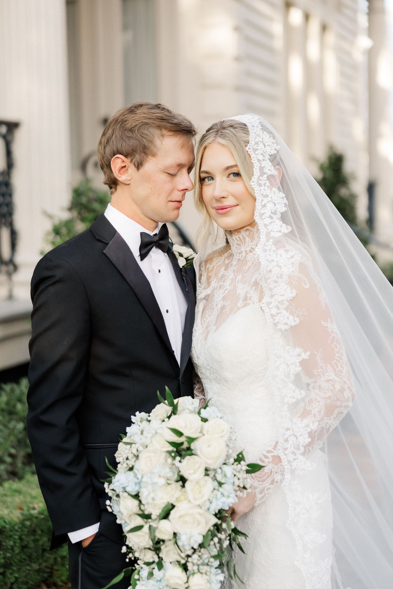Romantic wedding portrait against white columns with bride holding white floral bouquet.