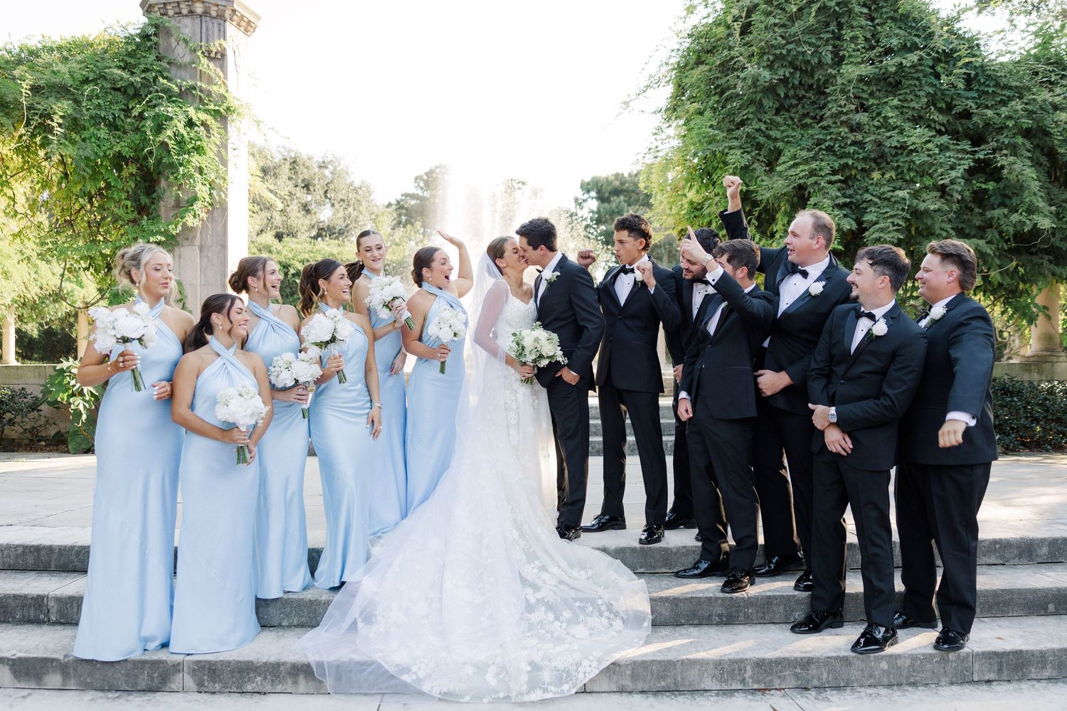 Wedding party in formal attire poses together on outdoor stone steps during summer celebration.
