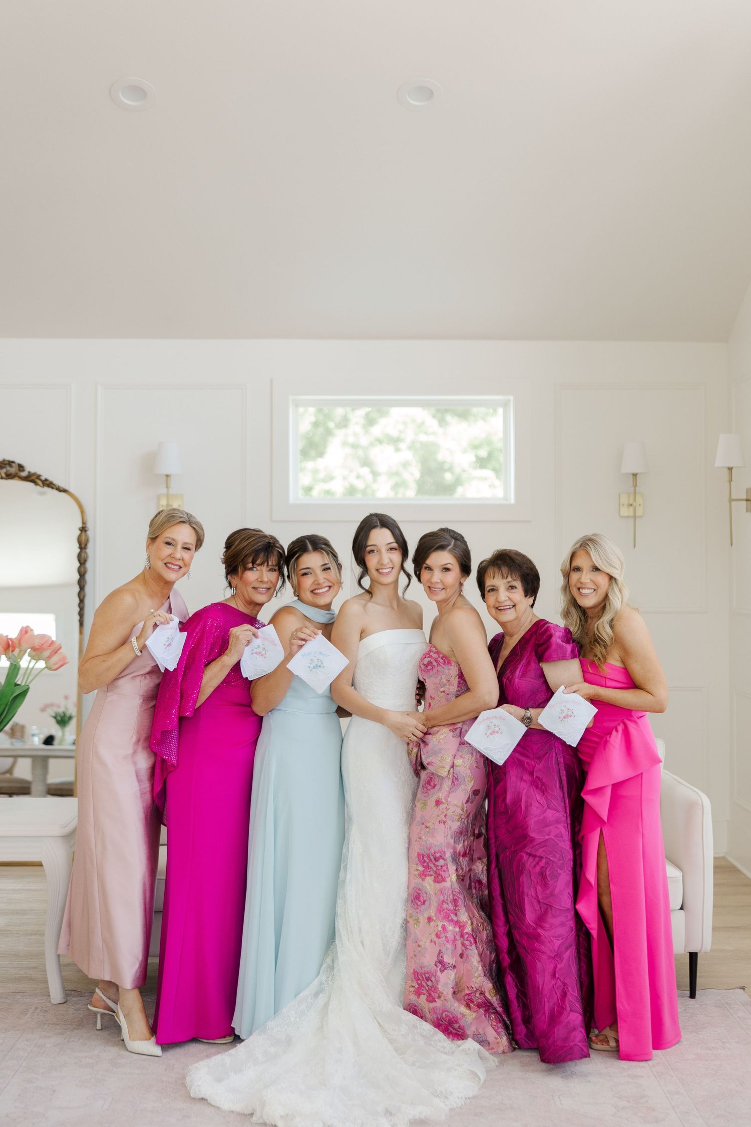 Bride in white gown poses with bridesmaids in mixed pink and blue dresses holding cards in bright room.
