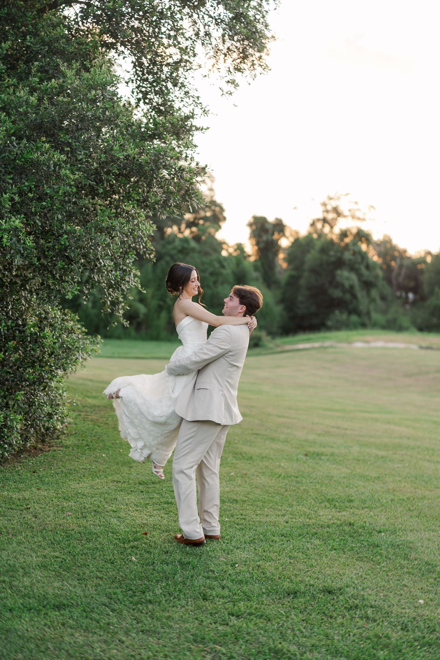 Groom lifting bride in white wedding dress on a lush green lawn at sunset during wedding portraits.