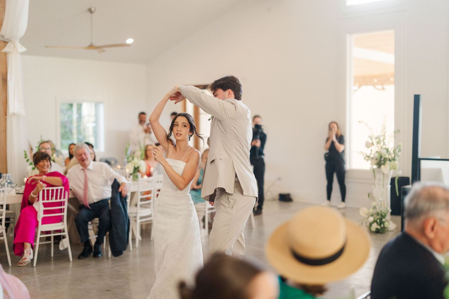 Bride and groom share their first dance in a bright white reception hall surrounded by seated wedding guests.