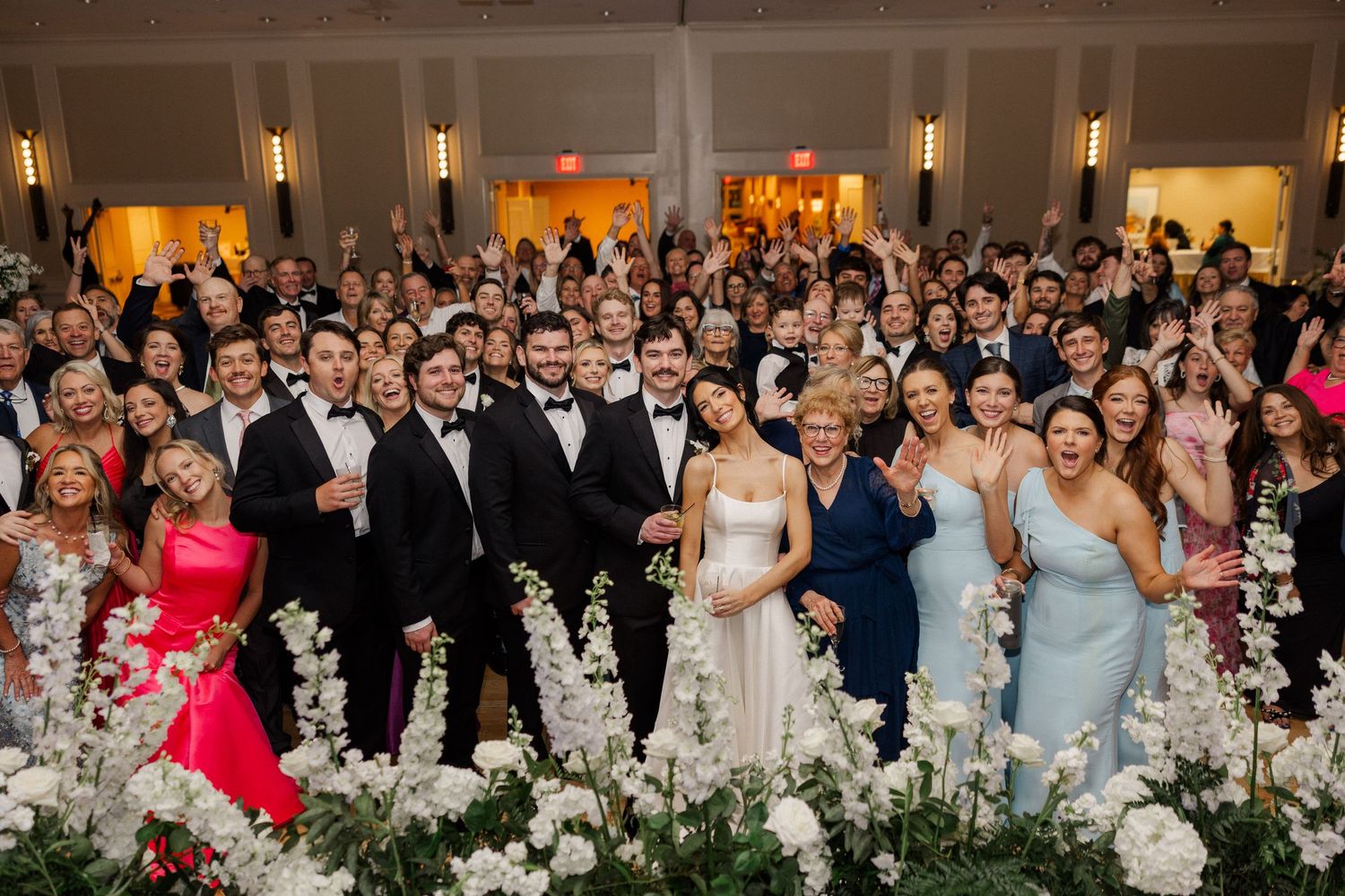 Large wedding group photo with guests celebrating at elegant white floral decorated reception venue.