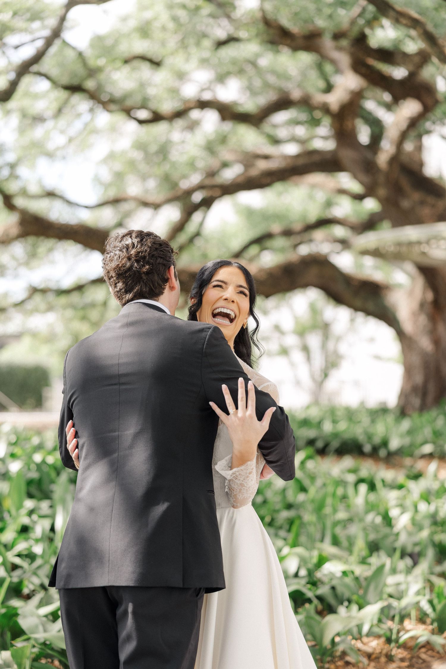 Joyful bride laughs and shows ring while embracing groom during outdoor first look.