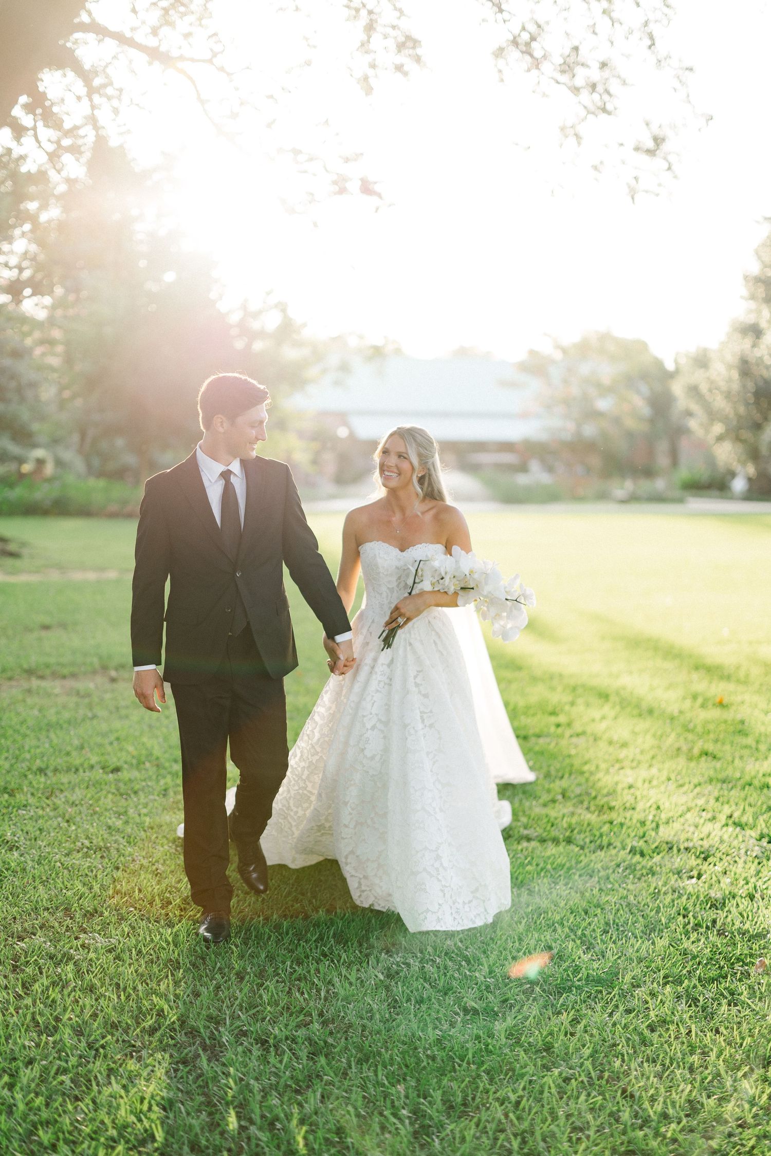 Bride in lace ball gown and groom in black suit walk hand in hand across green lawn in golden sunset light.