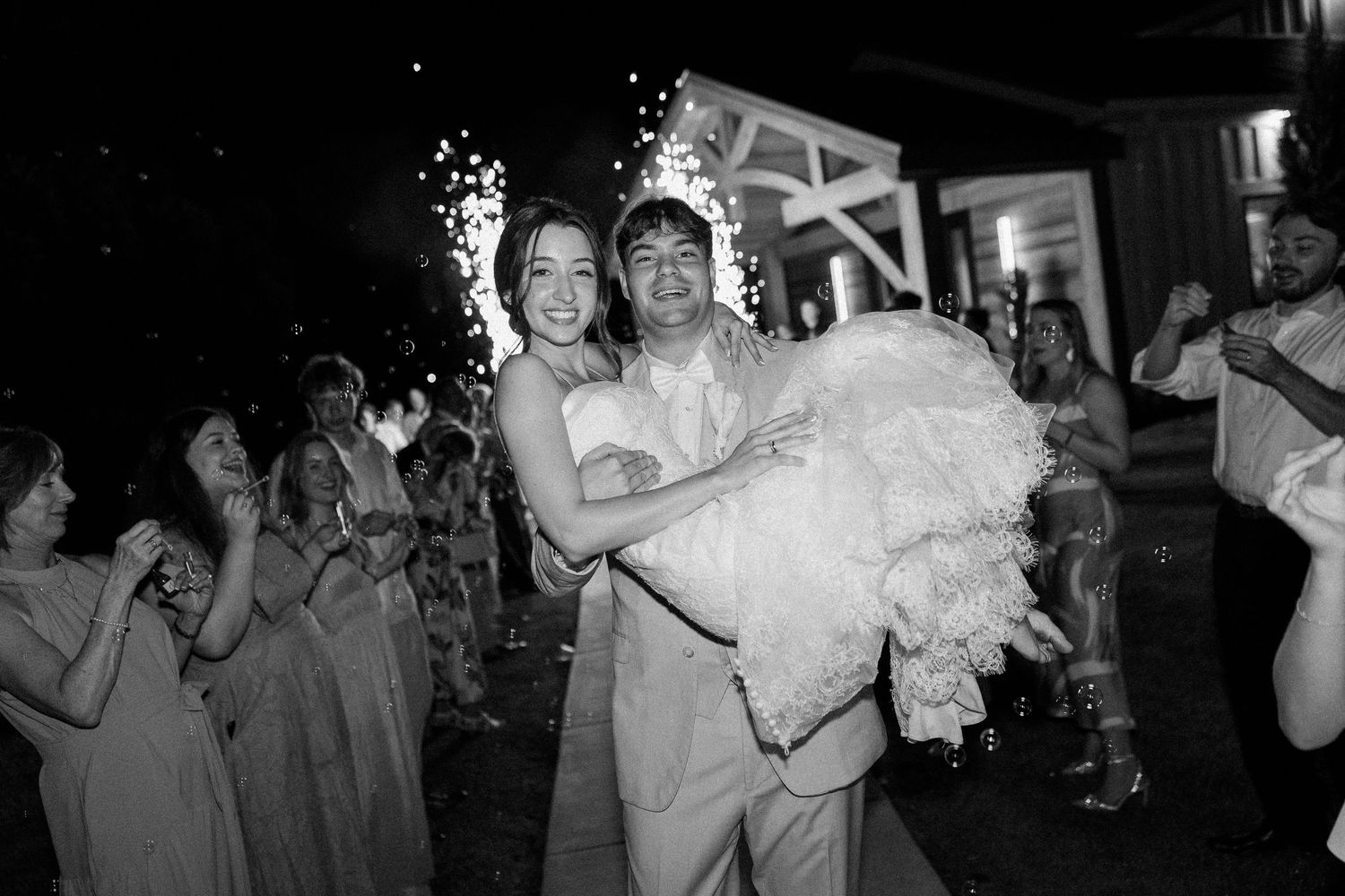 Black and white photo of groom carrying bride through cheering guests during wedding reception exit.