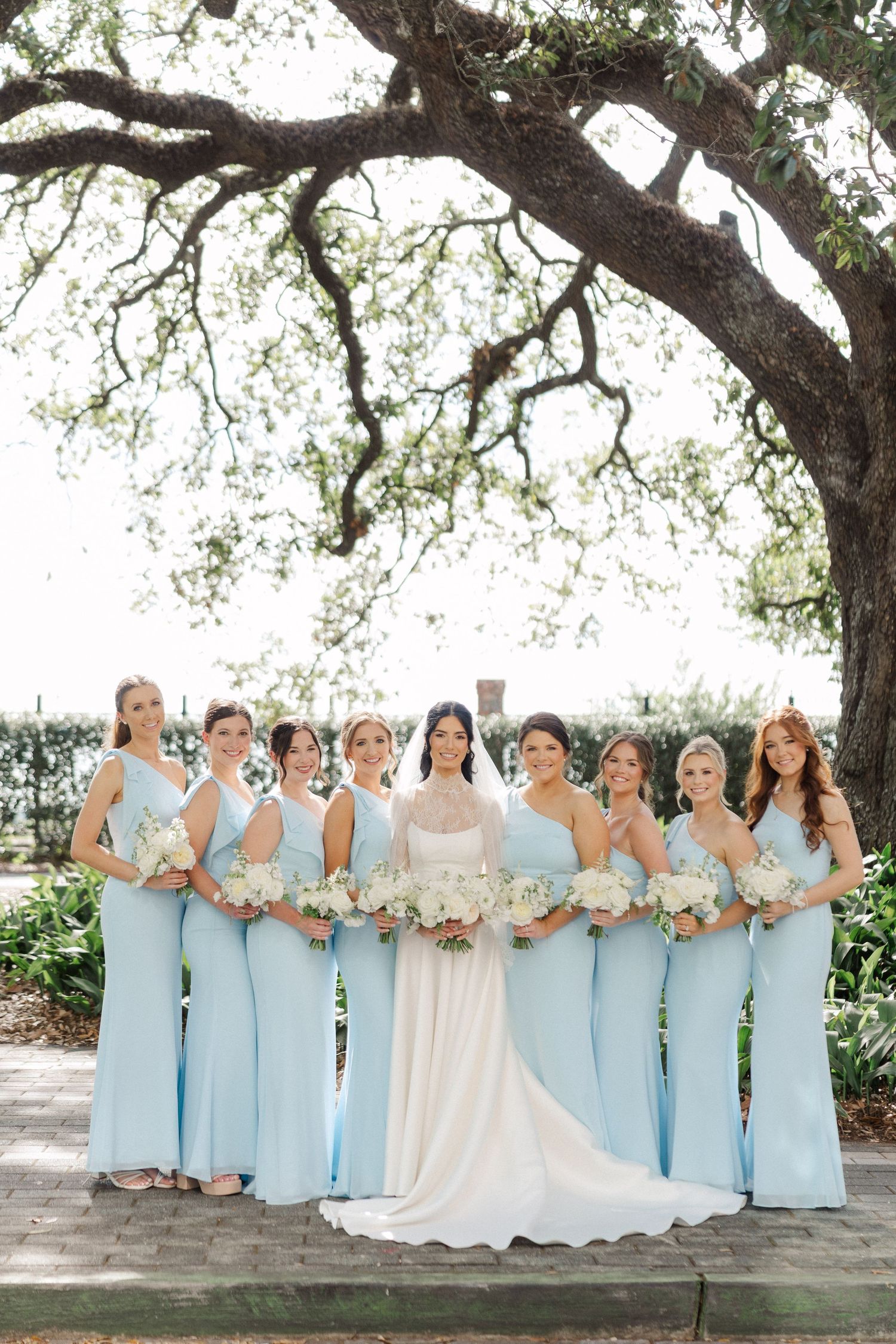 Bride in white gown poses with eight bridesmaids in light blue dresses holding white bouquets under large oak tree.