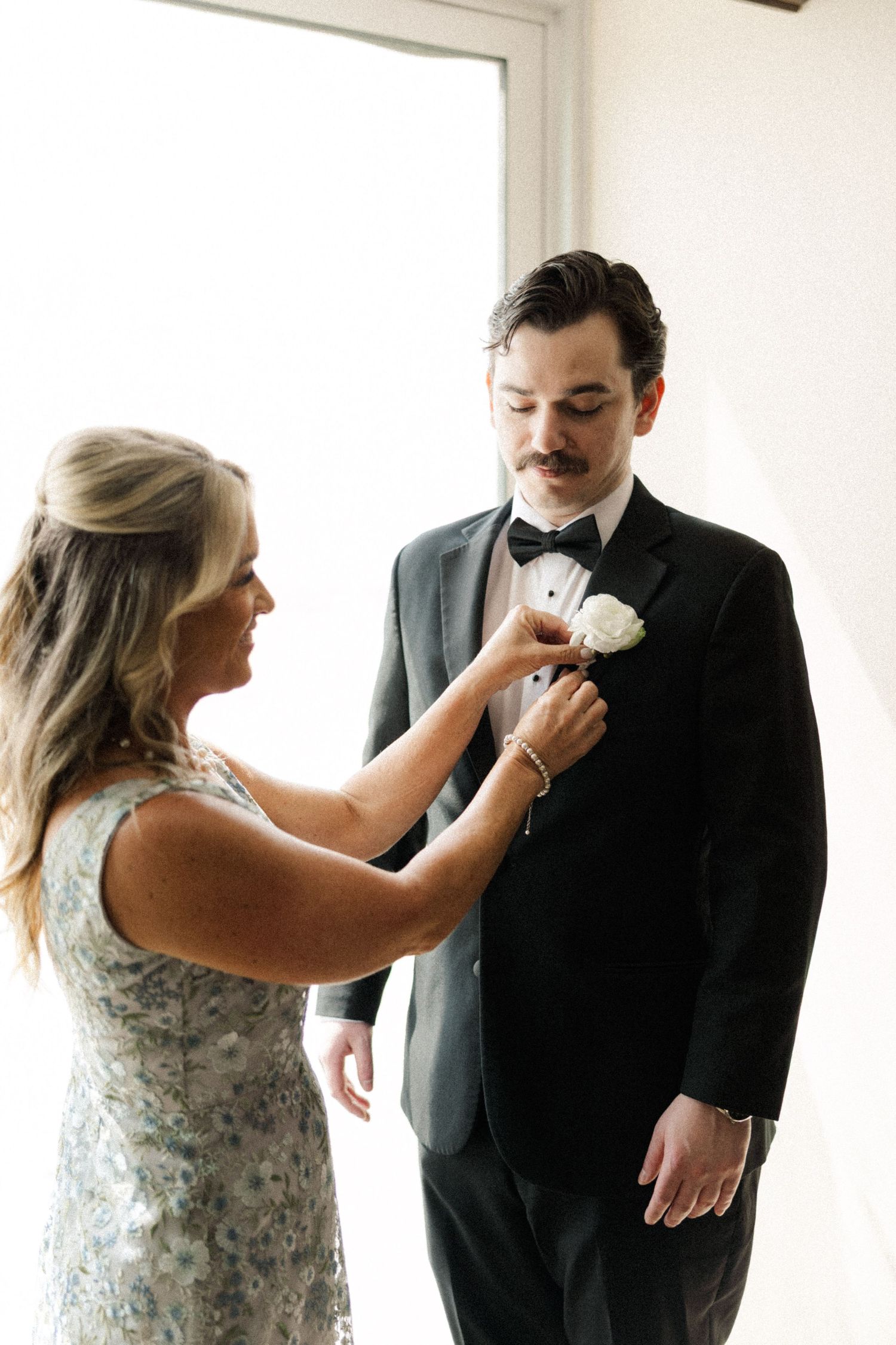 Mother pins white floral boutonniere on groom's black tuxedo lapel during wedding day preparations.