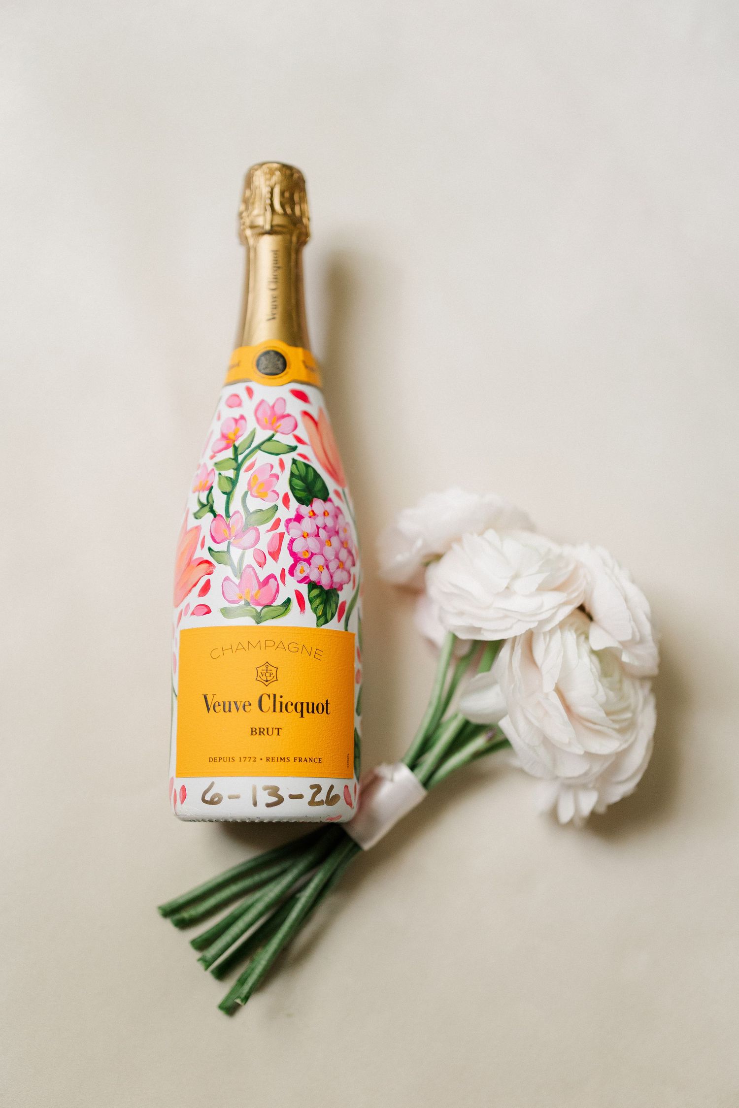 Veuve Clicquot Brut champagne bottle with floral painting and date 6-13-26, beside white ranunculus flowers.