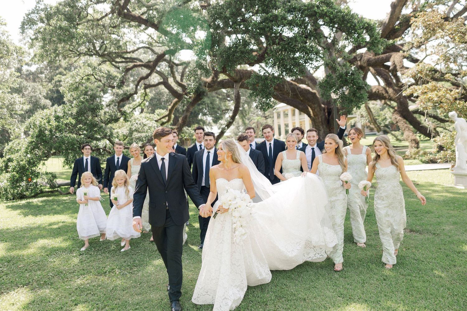 Bride and groom leading wedding party walk across green lawn under sprawling oak trees.
