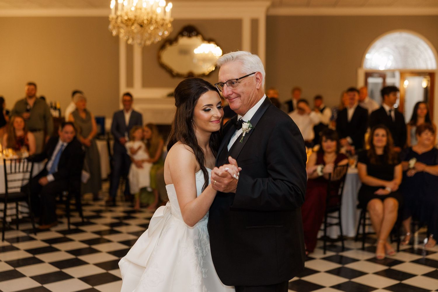 An emotional dance moment on a black and white checkered floor under a crystal chandelier at an elegant wedding reception.